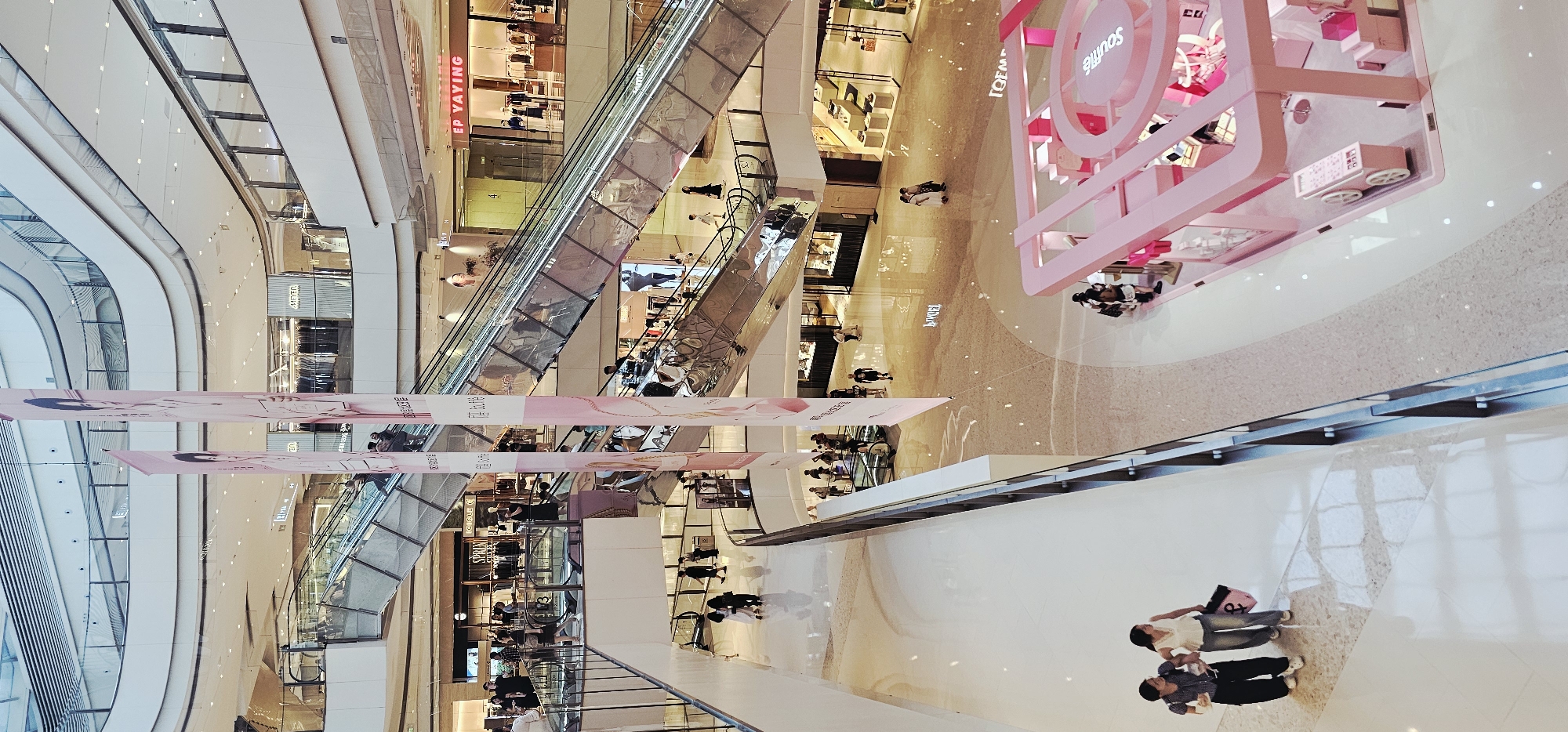 The image shows a multi-level interior of a modern shopping mall, likely located in Jiulongpo District, China, given the provided context. Escalators are visible, connecting different floors, with several people using them. Banners featuring portraits and text hang from the ceiling, adding to the commercial atmosphere. Various retail stores are open, displaying merchandise and mannequins. In the foreground, two women are walking on a polished floor, one carrying a shopping bag. Another area of the mall features a bright pink, contemporary retail display or kiosk, also with people nearby. The overall environment is bright, clean, and characterized by sleek architectural design and commercial activity.