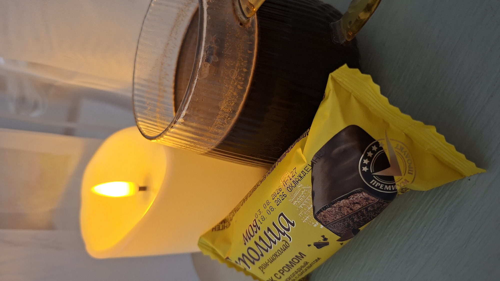 A close-up shot captures a cozy scene on a light-colored surface, likely a table. To the left, a large, unlit LED candle emits a warm, yellow glow, mimicking the appearance of a flickering flame. Beside it, a glass mug filled with dark coffee sits, its surface slightly frothy. In the foreground, a yellow wrapper of a chocolate-covered snack lies diagonally across the frame, displaying Russian text and an image of the treat. The packaging indicates it is a "Myatliya" brand chocolate and rum biscuit. The background features translucent white fabric, possibly curtains, diffusing the light. The overall ambiance is warm and inviting, suggesting a moment of relaxation. The image was taken in Myadzyel, Belarus.