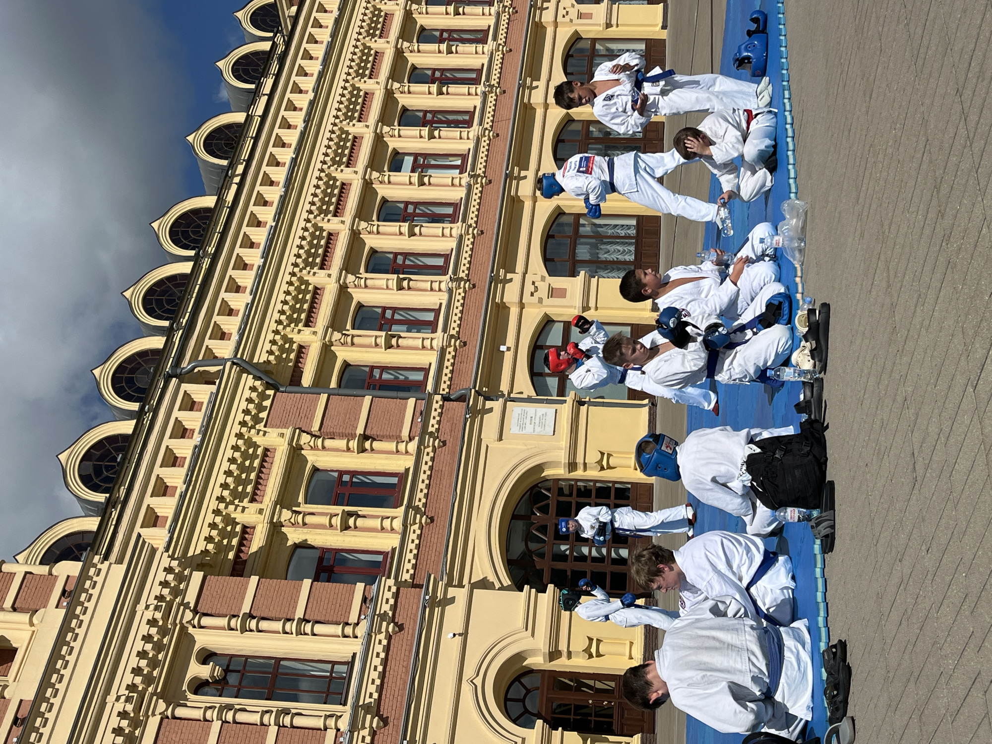 This image captures a group of young people, likely children and teenagers, practicing karate or a similar martial art on a blue mat in an outdoor urban setting. They are all wearing white gis and some have protective gear such as headguards and sparring gloves in blue, red, and black. The participants are in various stances, some standing and others sitting or kneeling. In the background, a large, ornate yellow and red building with arched windows and intricate architectural details is visible. This building is identifiable as the Nizhny Novgorod State Academic Opera and Ballet Theatre. The ground in front of the mat is paved with grey square tiles. Several water bottles and a black backpack are placed on the edge of the mat. The sky is blue with scattered white clouds, suggesting it is daytime. The overall scene depicts an organized outdoor training session for a martial art.