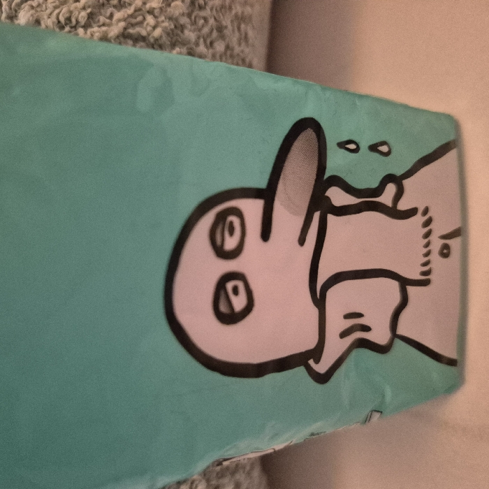 An image taken in Strasbourg, France, depicts a close-up of a cartoon character printed on a teal-colored flexible surface, likely packaging or a bag. The character features a light grey, rounded head with two large, tired-looking oval eyes, and a prominent, downward-pointing, beak-like nose. It wears a light grey garment with a ruffled collar and an outer cloak-like element. Two distinct teardrop shapes are depicted falling from the right side of the character's face, suggesting sadness. The teal surface shows slight creasing and is positioned against a textured, light grey/beige background on the upper right, and a smoother, lighter surface along the bottom edge.