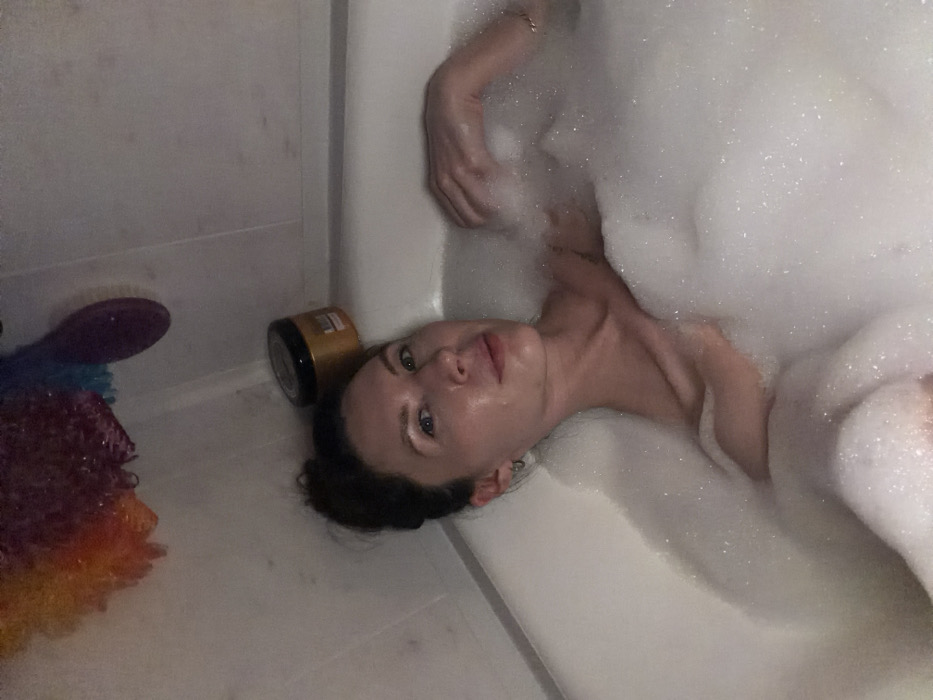 A single female individual is depicted partially submerged in a white bathtub filled with dense white foam. She maintains eye contact with the camera. Her dark hair is styled in an updo, and she wears small hoop earrings. Her upper torso and head are visible above the foam. On the rim of the bathtub, behind her head, rests a dark-colored jar with a gold-colored lid. Suspended from the white tiled wall above the tub are a multi-colored bath pouf and a dark purple, elongated bath accessory. The scene is set within a bathroom located in the city of Perm, Russia.