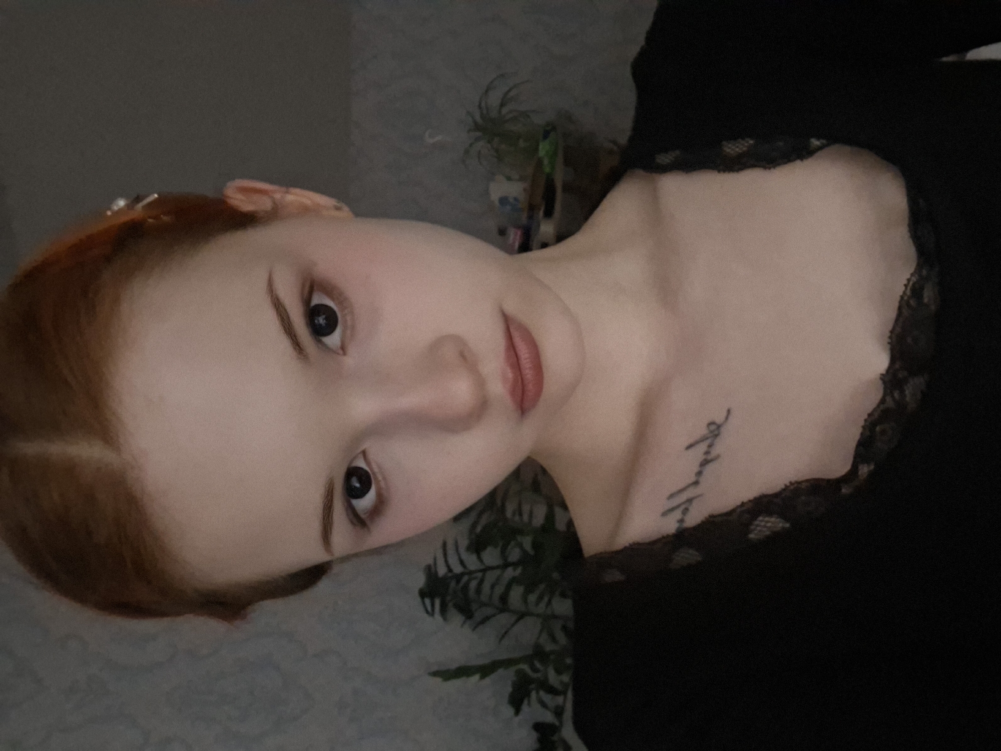 A close-up, rotated portrait shot depicts a fair-skinned young woman with reddish-brown hair pulled back, revealing a clear forehead. She has delicate features, including prominent cheekbones, dark eyes, and slightly parted lips with a neutral lipstick. She is wearing a black top with a lace neckline. On her upper chest, there is a small, cursive tattoo. The background is indistinct, showing a patterned surface and some greenery. The photograph appears to have been taken indoors. There is no information on the location of Chaikovsky, Russia to be determined from the photograph.