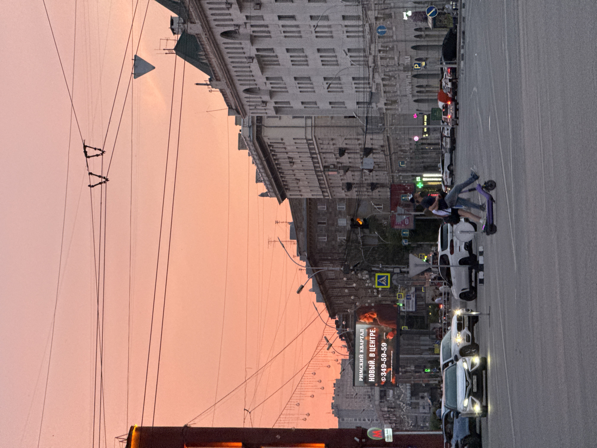 The image depicts a street scene in Novosibirsk, Russia, during sunset. The sky is a vibrant orange-pink, with power lines crisscrossing the frame. In the foreground, a street with multiple lanes of traffic is visible. Several cars are present, including a white Toyota Camry with its headlights on, a white BMW SUV, and other vehicles. A couple is seen in the middle of the street, with one person riding a purple electric scooter and the other walking alongside, holding them.
In the background, several multi-story buildings line the street. One building has a large digital billboard displaying an advertisement in Russian text that reads "РИМСКИЙ КВАРТАЛ НОВЫЙ. В ЦЕНТРЕ." (Rimsky Kvartal Novy. V Tsentre.) followed by a phone number. Traffic lights and street lamps are also visible. The overall impression is of a busy urban environment transitioning from day to night.