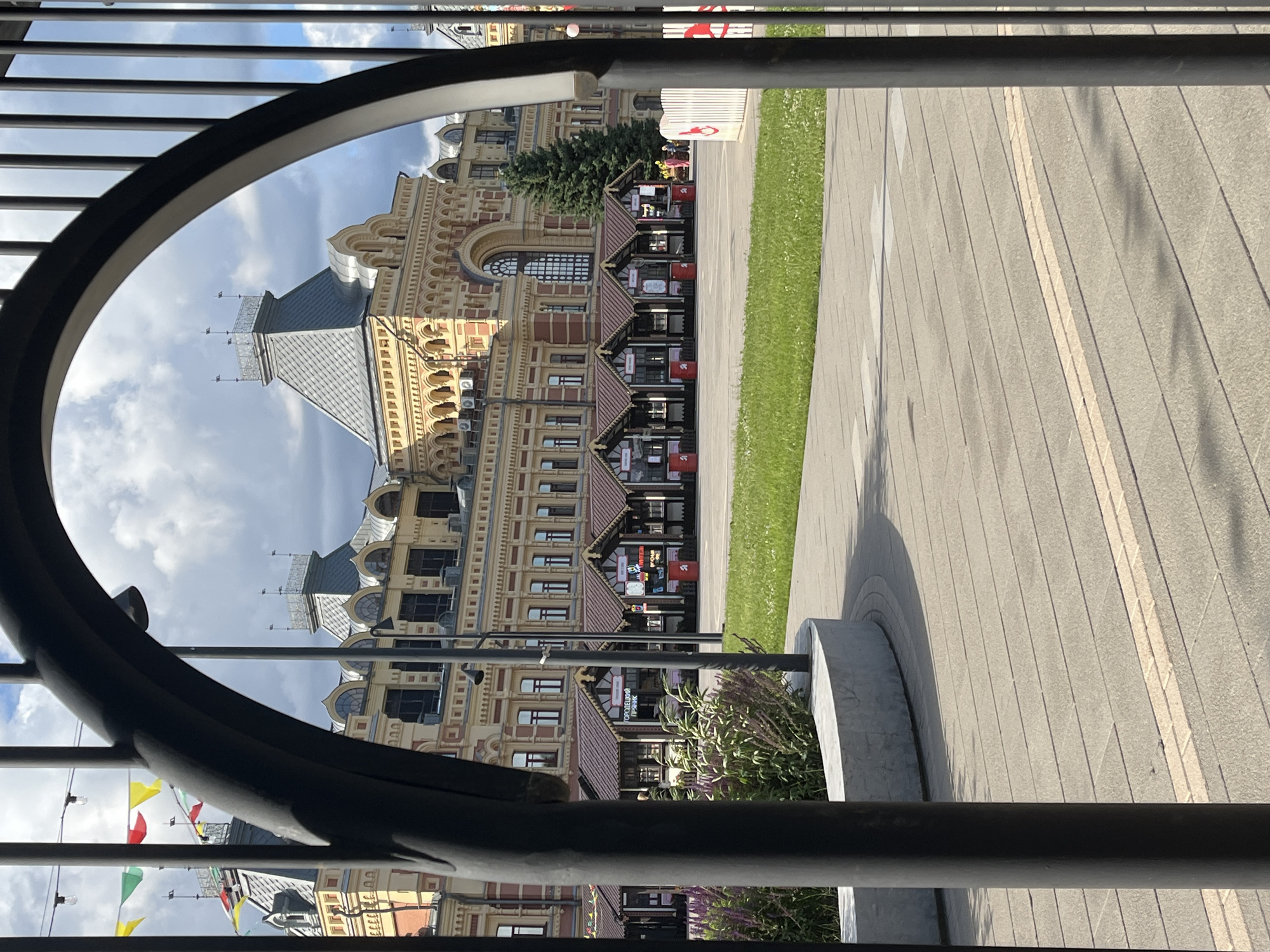 The image depicts a view through a large arched gate of the Nizhny Novgorod Fair (Nizhegorodskaya Yarmarka) in Nizhny Novgorod, Russia. In the foreground, a dark, curved metal arch and vertical bars of the gate are prominent, framing the scene. Beyond the gate, a large, ornate building with yellow walls, red accents, and a distinctive grey roof with turrets dominates the background. In front of this building, a row of small, pitched-roof market stalls with dark brown roofs are set up. Green grass covers the ground in the middle ground, leading to a paved plaza. A curved, grey stone planter with some green foliage is visible on the left side. The sky above is partly cloudy. A few people are visible walking in the distance near the market stalls.