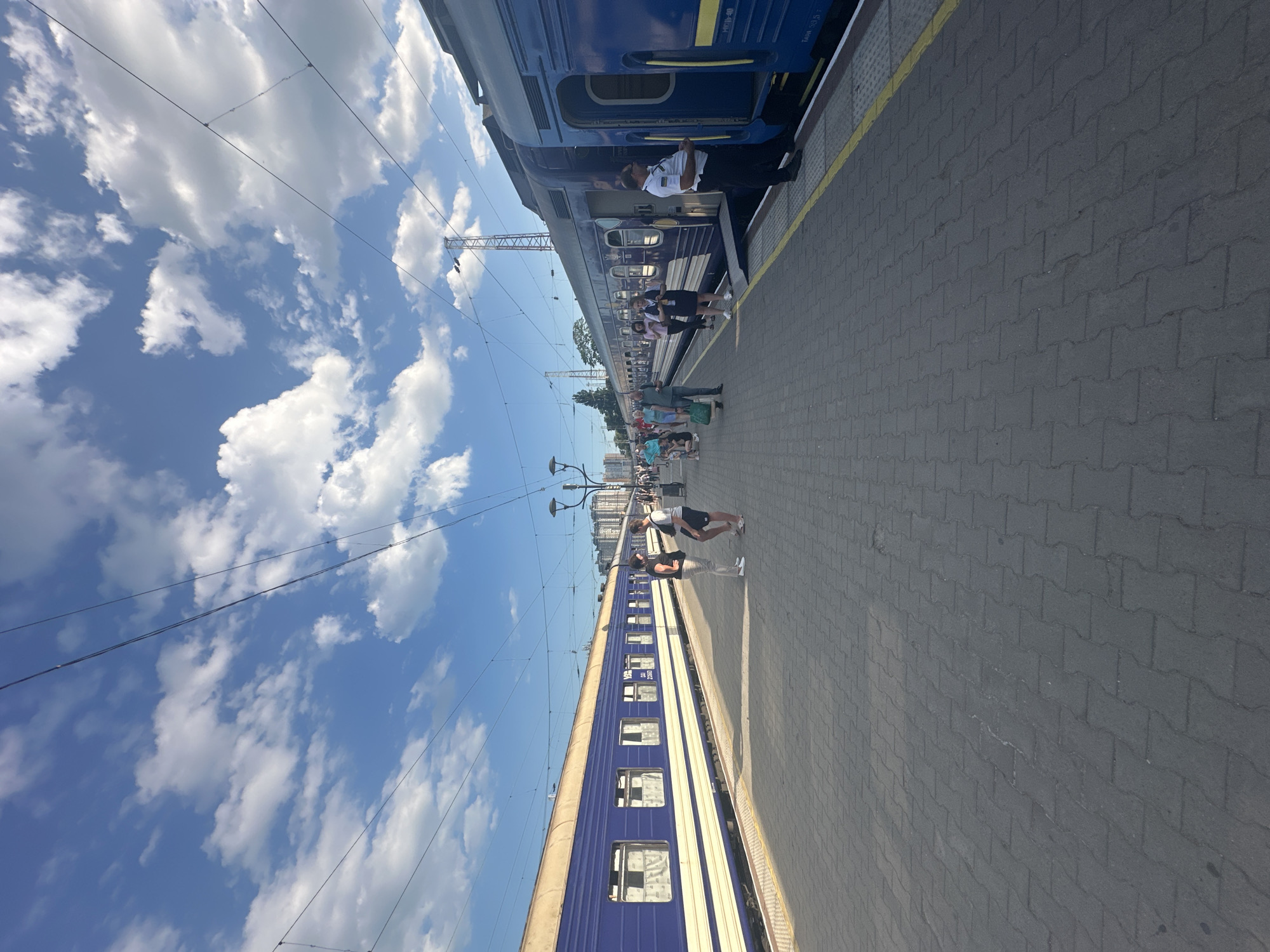 The image depicts a train station platform in Odesa, Ukraine, on a clear day with a bright blue sky filled with white cumulus clouds. Two blue and yellow passenger trains are stopped at the platform, one on the left and another on the right. The train on the left is longer and appears to be parked parallel to the platform. The train on the right is positioned at an angle, with its front part extending further into the frame and its doors open. Several passengers are visible on the platform, some standing and conversing, while others are in queues or walking. A train conductor in uniform is standing by the open door of the train on the right. The platform itself is paved with interlocking gray bricks and has a yellow line marking the edge. In the background, buildings and infrastructure typical of a city are visible, along with overhead electrical wires for the trains and streetlights.