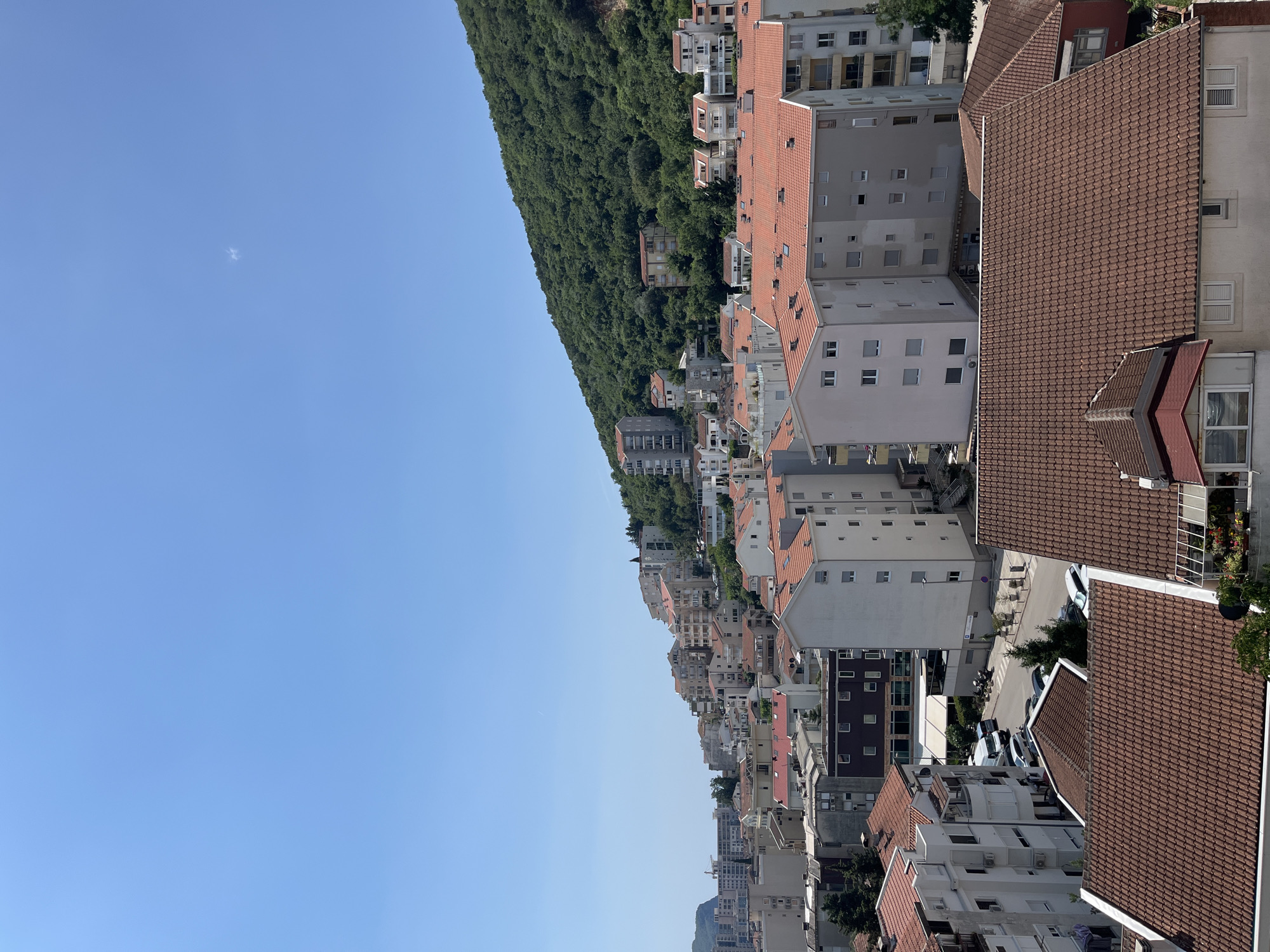 This is an aerial, high-angle, medium shot of Budva, Montenegro, on a clear, sunny day. The image captures a dense urban landscape dominated by apartment buildings with terracotta tile roofs, nestled against the base of a lush, tree-covered mountain. The buildings are of varying heights and architectural styles, primarily white and light-colored, with many balconies and windows. In the foreground, a large apartment building with a red tile roof is prominently visible. Below this, a street with parked cars and a white van can be seen. The mountain slopes upward to the right, covered in dense green foliage. The sky is a clear, bright blue with a few wispy clouds and a faint contrail. The scene suggests a coastal city with a Mediterranean climate, characterized by its distinct architecture and natural surroundings.