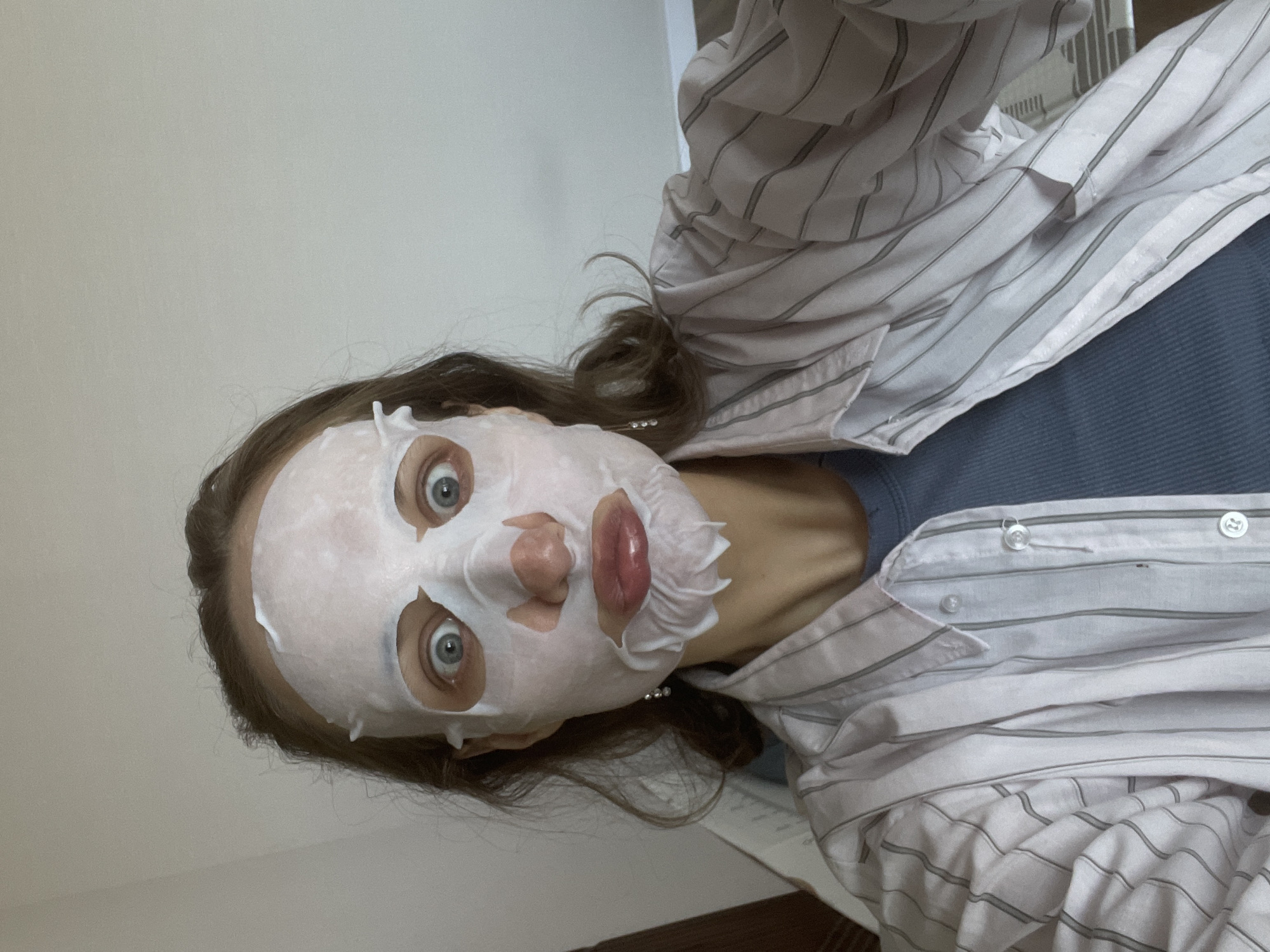 A woman is seen from a close, tilted, high-angle perspective. She is wearing a sheet mask, which is a white, non-woven fabric infused with serum, fitted over her face with cutouts for the eyes and nostrils. Her eyes are visible, and her lips protrude through the mask. She is wearing a blue top underneath a white and grey striped collared shirt, which is partially unbuttoned. Her brown hair is pulled back. The background consists of a light beige wall, suggesting an indoor setting. While it's not possible to determine the precise location as Sepych, Russia with the information provided, the scene depicts a person in what appears to be a home or residential environment wearing a face mask.
