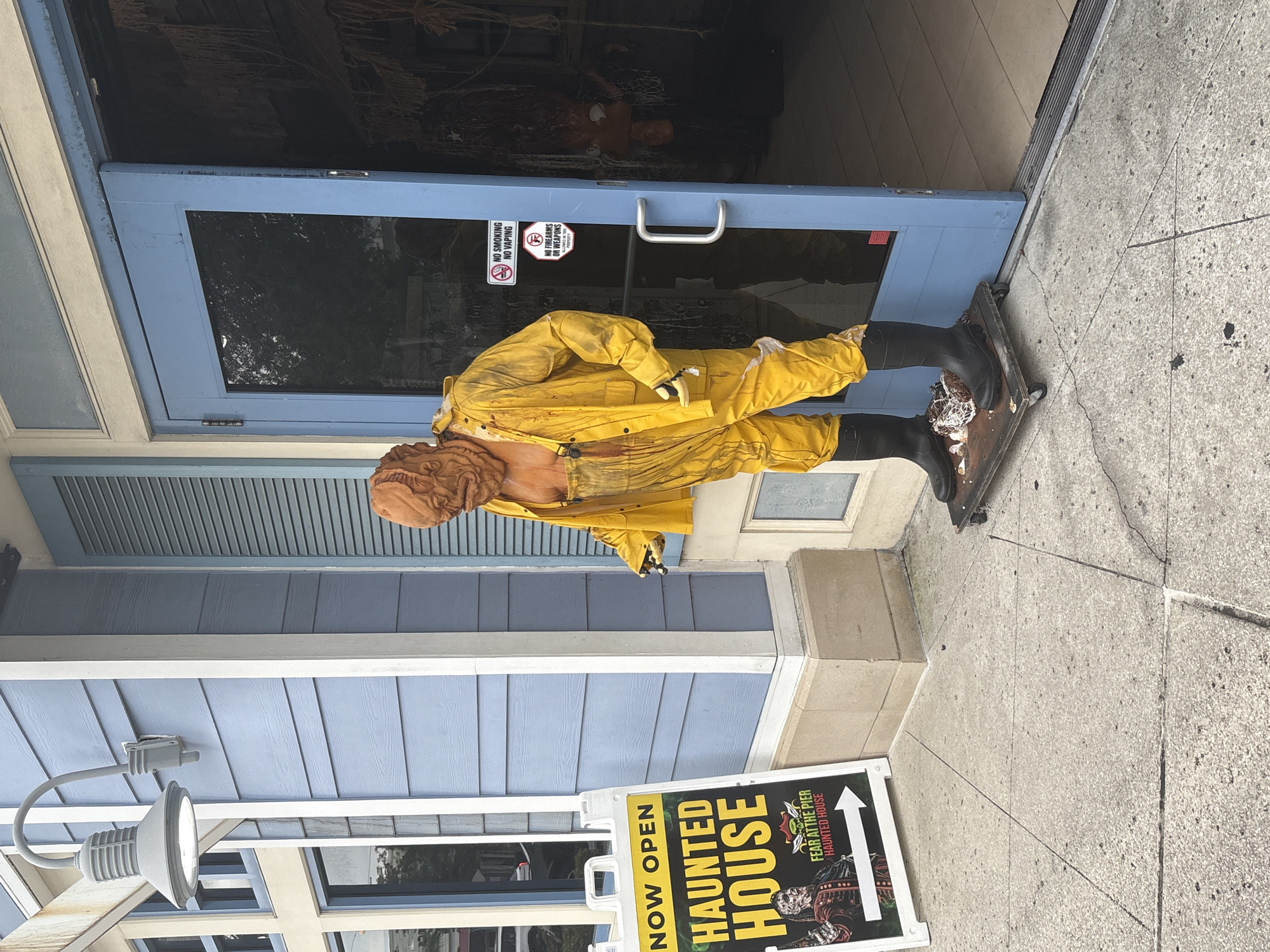 An outdoor, eye-level, medium shot captures a spooky scene in front of a haunted house attraction in Panama City Beach, Florida. A large, grotesque figure, clad in a yellow slicker and overalls, stands on a wheeled platform just outside a blue double door. The figure's skin is a rough, brown texture, and its face is contorted in a menacing expression. Bloody splatters adorn its clothing, adding to the eerie ambiance.
To the left of the figure, a white sign with bold black and yellow lettering announces "NOW OPEN HAUNTED HOUSE," with a smaller subtitle reading "FEAR AT THE PIER HAUNTED HOUSE." The building behind the figure features light blue siding and a light fixture. Two "No Smoking" and "No Firearms or Weapons" signs are affixed to the blue door. The ground is a concrete sidewalk with visible cracks. The scene suggests a commercial establishment designed to evoke fear and excitement.