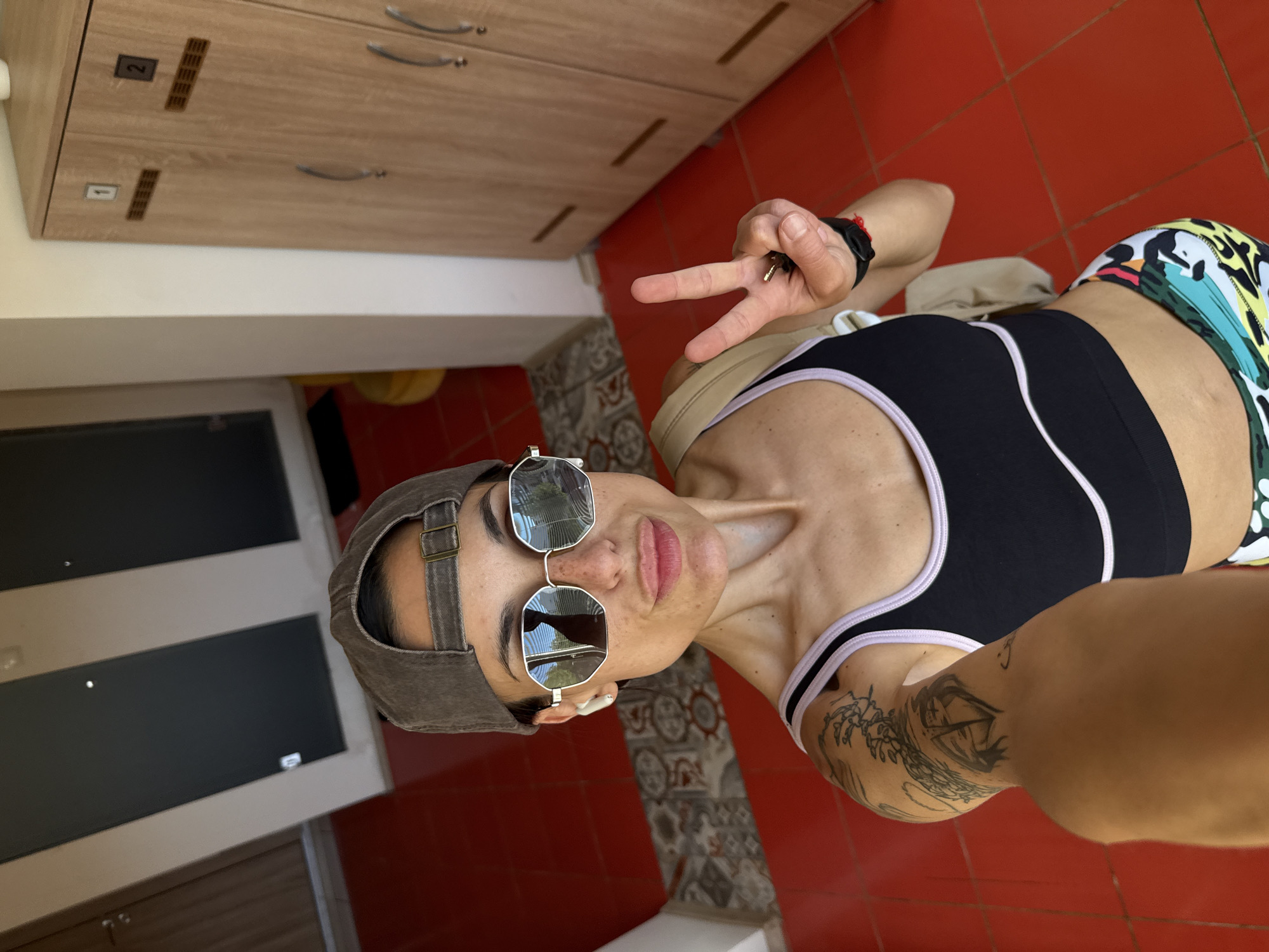 A person, likely female, is taking a selfie in what appears to be a locker room. The person is wearing a baseball cap, sunglasses, a black sports bra with light pink trim, and patterned shorts. They have tattoos on their left arm and are holding up two fingers in a peace sign. The locker room has red tiled floors and light wood lockers with numbers 1 and 2 visible. The exact location in Pleven, Bulgaria cannot be determined from the image.