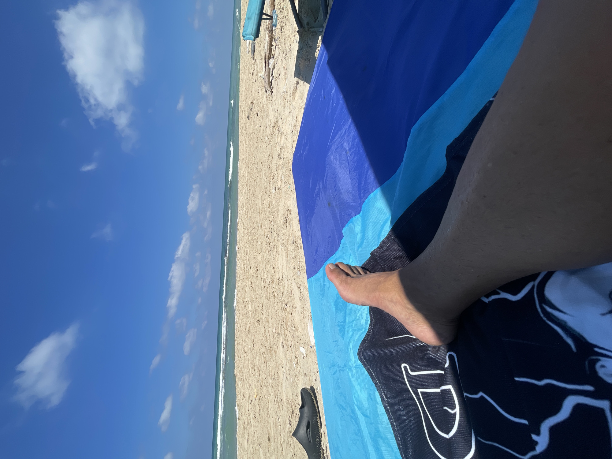 In Ga'ash, Israel, a person is relaxing on a beach. The image is taken from a first-person perspective, focusing on the person's legs and feet as they lie on a towel. The towel is blue and black with a graphic design. Next to the towel are a black sandal and some sand. In the background, the sea and the sky are visible. The sea is a vibrant blue-green with white waves, and the sky is a clear blue with a few scattered white clouds. A beach umbrella and some driftwood are also present in the background. The overall scene depicts a tranquil day at the beach.