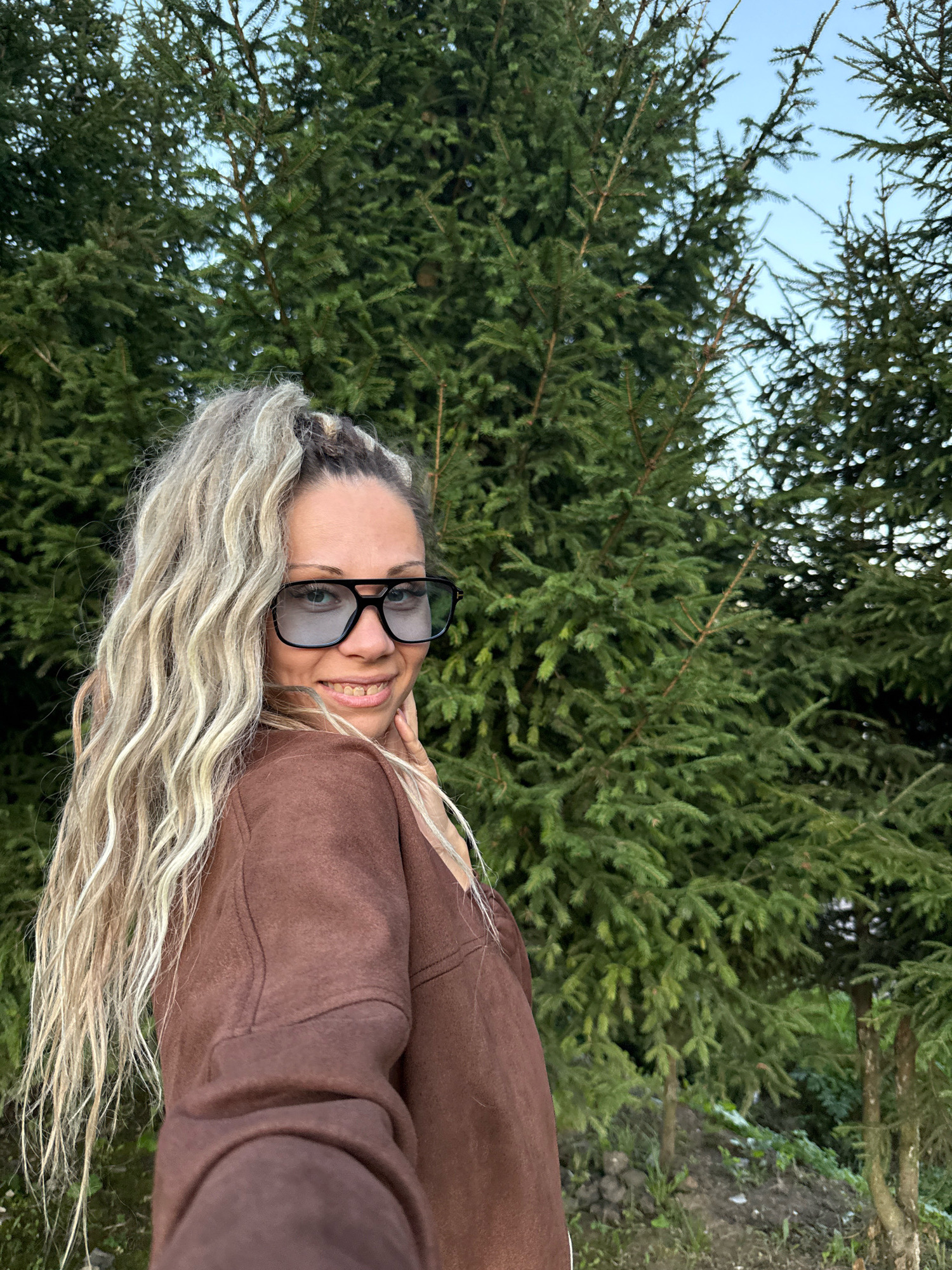 A medium shot shows a fair-skinned woman from the chest up, looking over her right shoulder at the camera. Her wavy, blonde hair is pulled back into a ponytail. She wears dark-rimmed glasses and a brown sweatshirt. The woman is positioned in front of a backdrop of tall, dark green pine trees. The lighting suggests it is daytime.