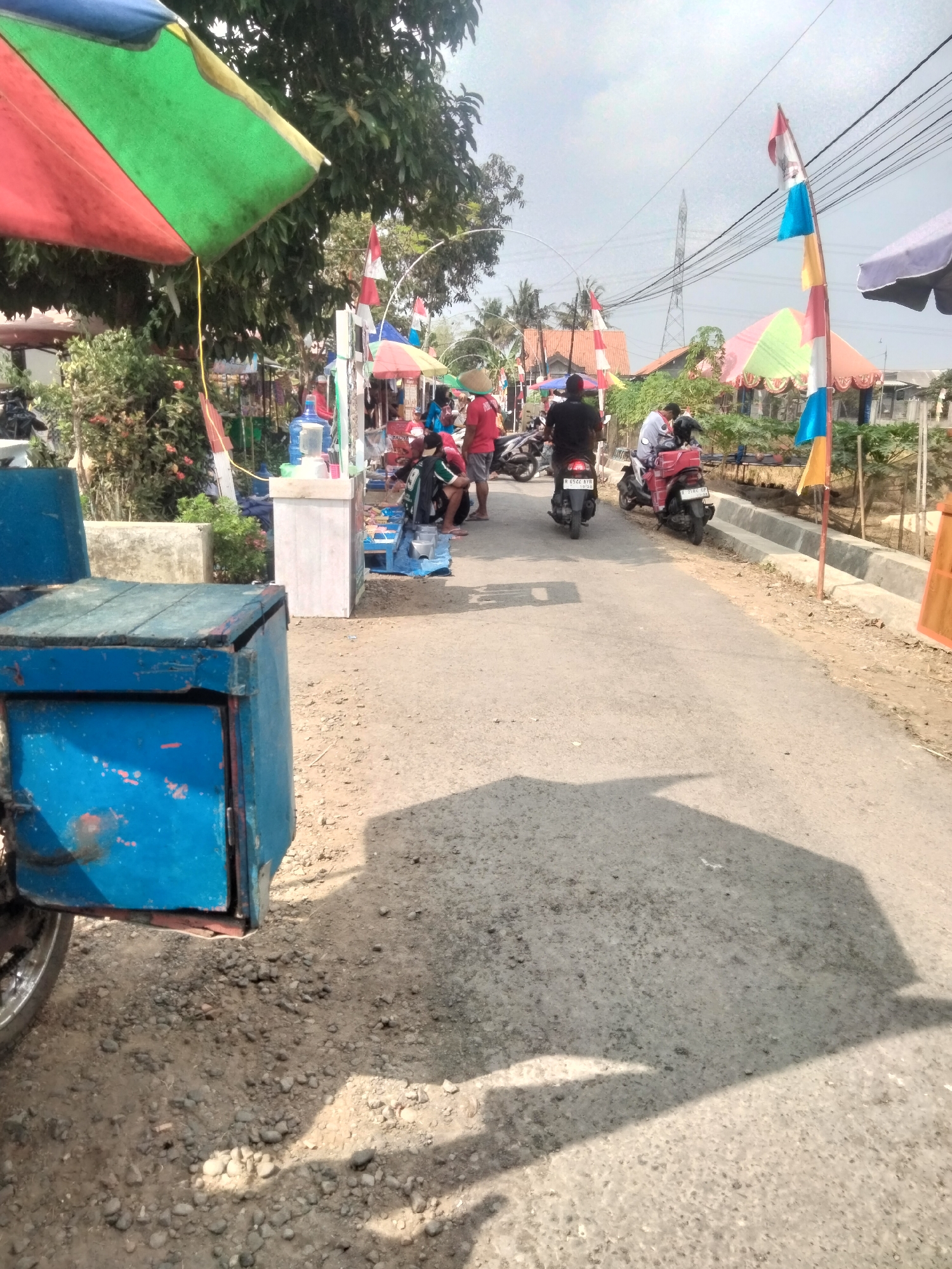 In Ketanggung, Indonesia, a paved road runs through a marketplace with various stalls and vendors. Several people are seen interacting with vendors and riding motorcycles. Colorful umbrellas and flags adorn the scene, indicating a festive atmosphere. The foreground features a weathered blue cart. The sky is overcast, and the overall environment suggests a warm, humid climate.