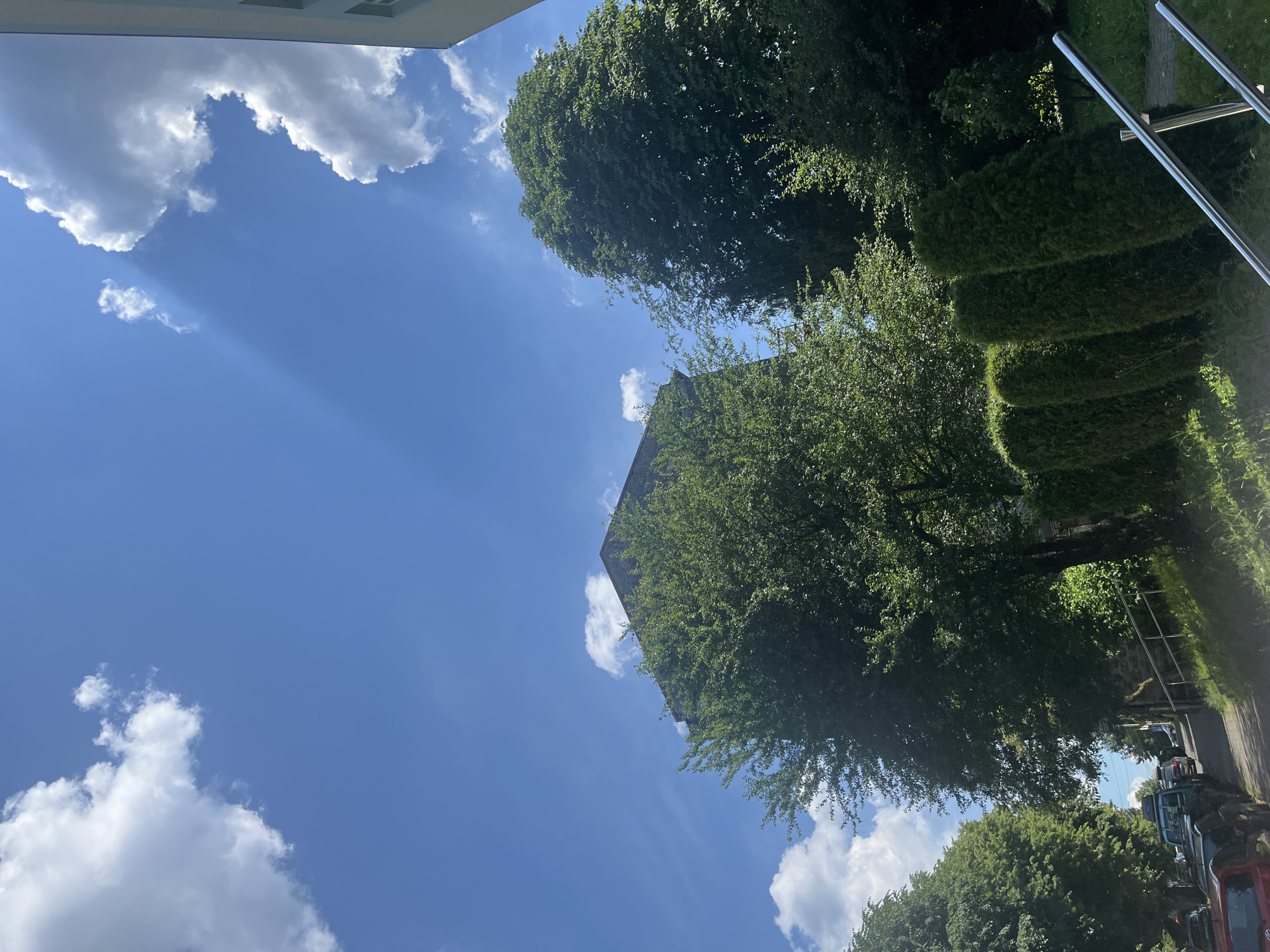 The media file shows an outdoor scene with a bright blue sky and fluffy white clouds. The image is oriented vertically, with the sky occupying the majority of the frame. Sunlight is streaming through the clouds, creating visible rays of light.
In the foreground and middle ground, there are lush green trees with dense foliage, and a well-maintained, stepped hedge. A portion of a building with a light-colored facade is visible at the top of the image. In the background, a street with parked cars can be seen, suggesting an urban or suburban setting. The trees are so abundant that they partially obscure the view of the sky.
The scene appears to be set during the daytime, given the bright sunlight and clear blue sky. The weather seems pleasant and sunny. There is no discernible text in the image. The overall impression is one of a serene, natural environment contrasted with signs of human habitation. The image is a low-angle shot, looking up towards the sky and the surrounding elements.