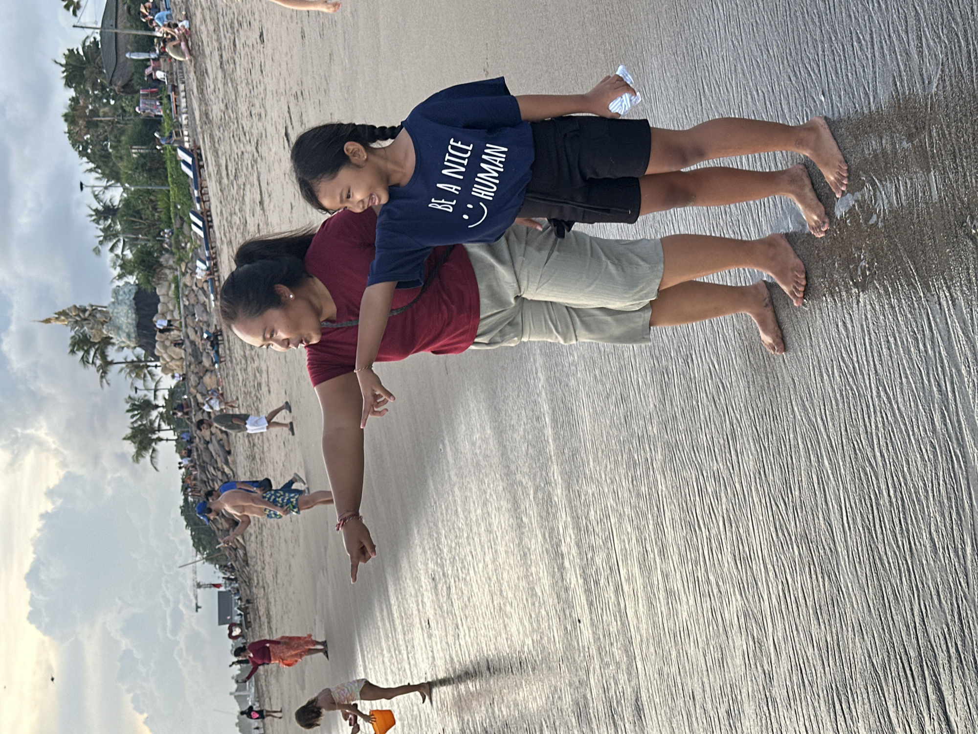 The image shows a woman and child standing on a beach with wet sand. The child is wearing a navy blue t-shirt that says, "BE A NICE HUMAN." The woman is wearing a maroon shirt and light-colored shorts. She is pointing towards the beach. Other people are present on the beach in the background. Palm trees are visible near a building in the background. The location is North Kuta, Indonesia.