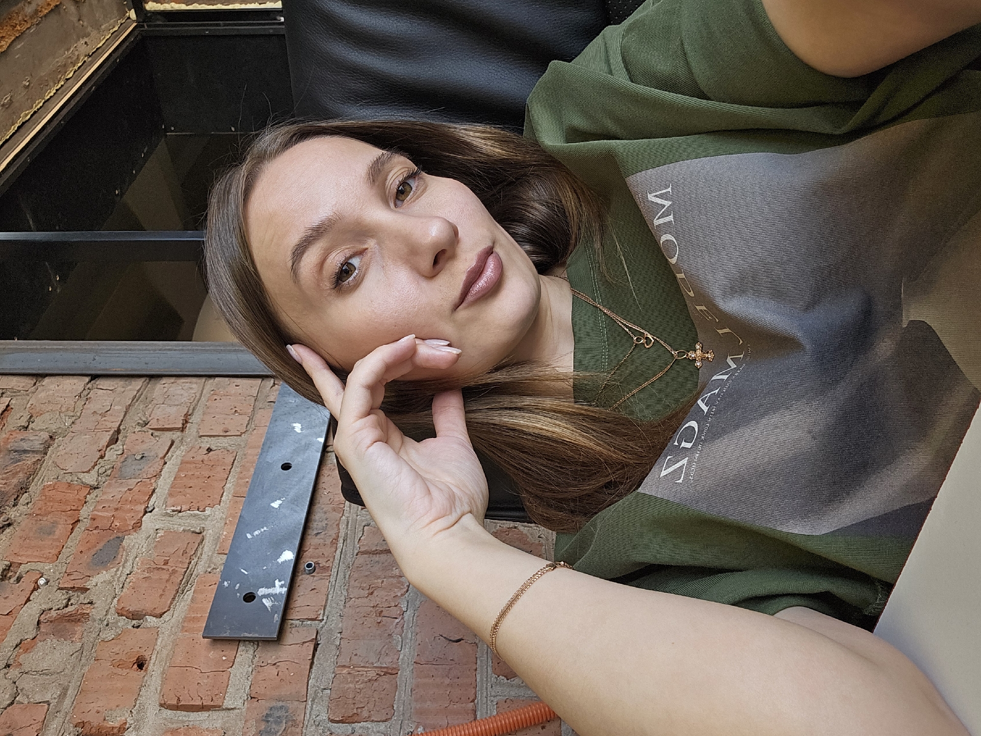 A woman poses for a selfie. She has brown hair, fair skin, and is wearing an olive-green shirt with gray accents and gold jewelry. Her left hand is touching her face. The background consists of a brick wall, a black metal fixture, and what appears to be part of a window or door frame. The location is Chaikovsky, Russia.