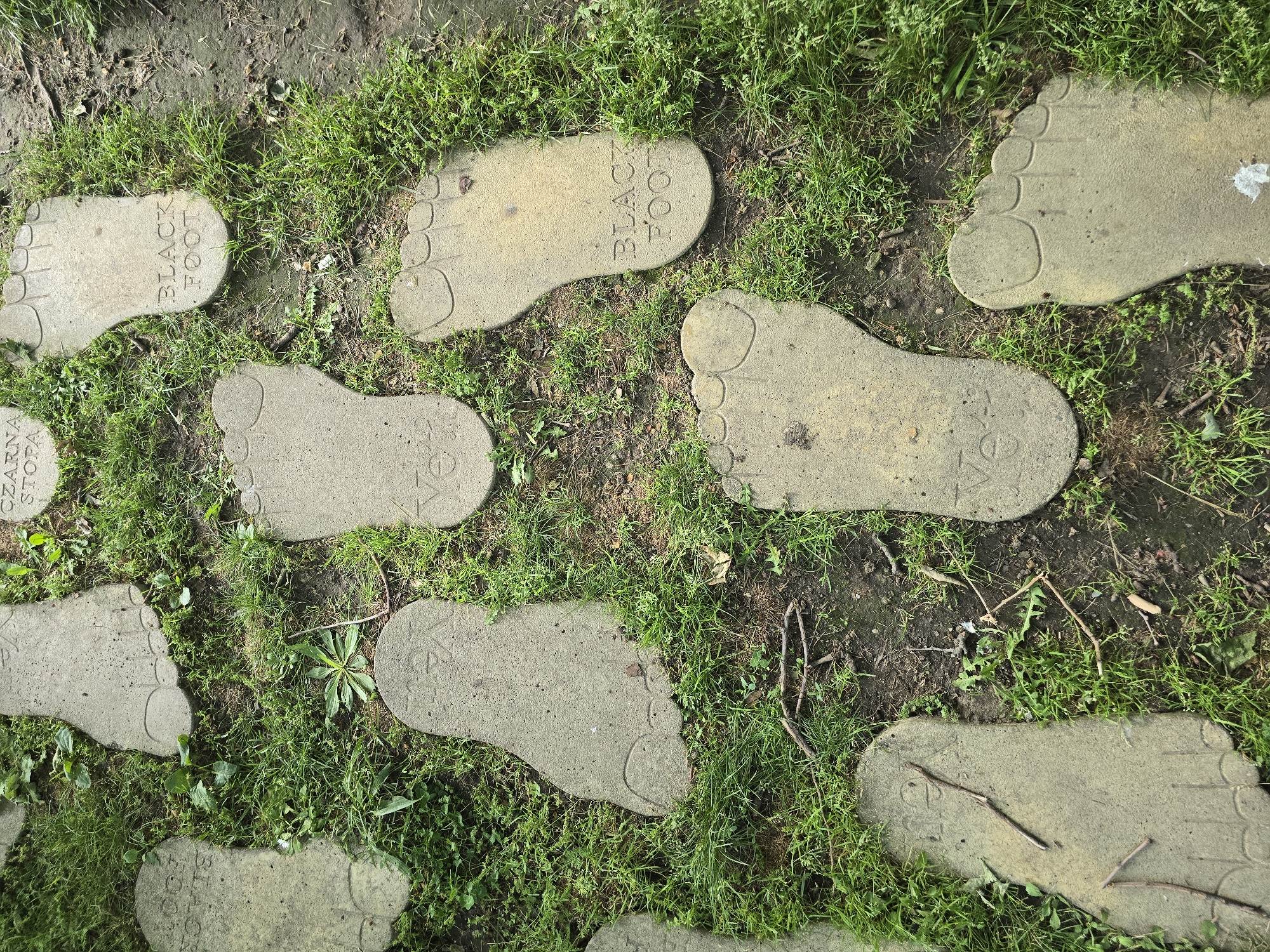 The image shows a path made of concrete footprints set in grass and dirt. Some footprints are labeled "BLACK FOOT" while others have "CZARNA STOPA" inscribed on them. The location is not identifiable.