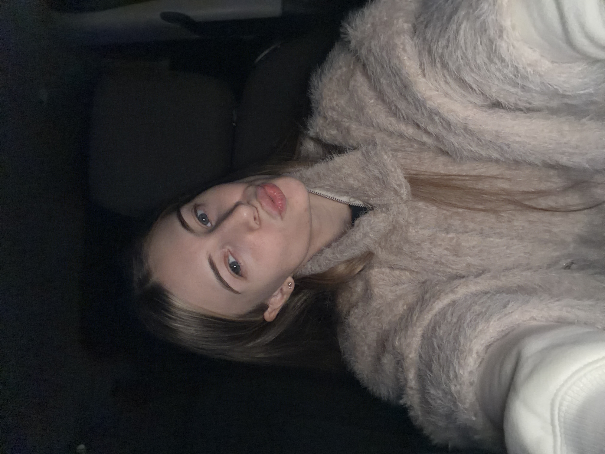 A young woman with light brown hair and blue eyes is in the driver's seat of a car. She is wearing a fuzzy, light brown jacket and a white sweatshirt. The car interior is dark. The location cannot be determined as Plovdiv, Bulgaria, from the available information.