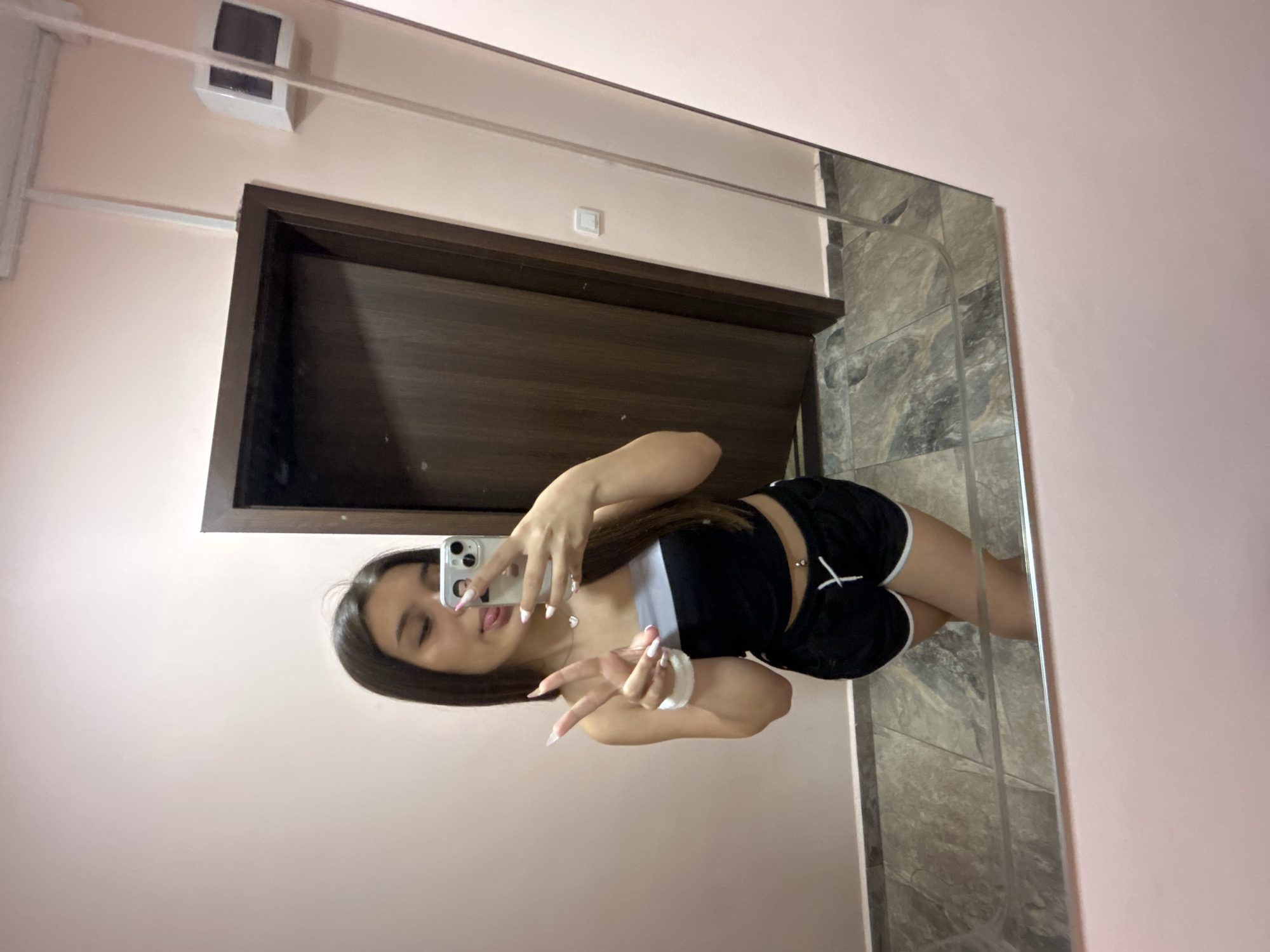 A young woman is taking a selfie in a mirror. She is wearing a black strapless top and black shorts with white trim. She has her tongue sticking out and is making a peace sign with her fingers. The background is a pink wall and a dark wood door. The floor is tiled. There is a light switch on the wall next to the door and a security camera on the wall above the door. The scene is indoors, likely in an apartment or a building. This location is in Shumen, Bulgaria.