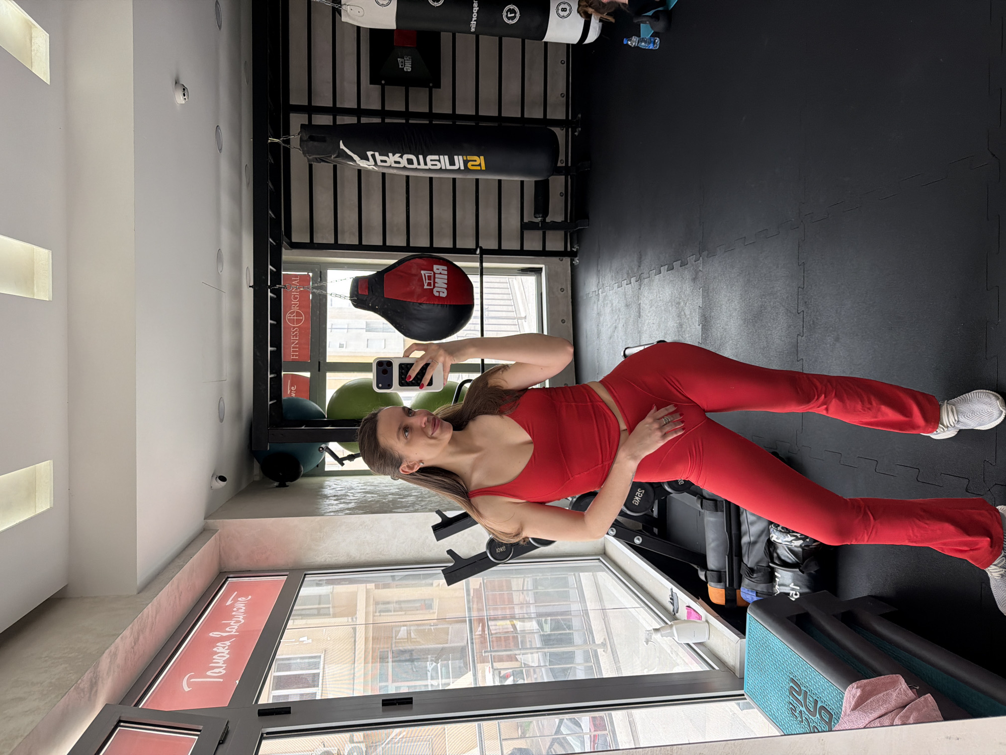 An interior shot depicts a female in a fitness gym, capturing a mirror selfie. The woman, with light brown hair partially pulled back, wears a red two-piece athletic outfit consisting of a cropped V-neck top and flared pants, complemented by white and grey athletic shoes. She holds a smartphone with a patterned case in her right hand, angled to photograph her reflection.
The gym features black interlocking rubber flooring and large windows along the left wall, offering a view of an urban street with multi-story buildings. Directly outside the window, a red banner displays the white text "Tamara Radinović." Inside, a sign above punching bags reads "FITNESS ORIGINAL." Gym equipment includes a red and black teardrop-shaped punching bag, two black cylindrical punching bags (one with yellow and white "SPORTRING" text), a dumbbell rack, a blue and black exercise step platform with a pink towel, and a black kettlebell with a water bottle on a teal mat in the background. A wall-mounted pull-up bar or ladder structure is also present. A white spray bottle and a pink cloth are visible on the window ledge. The ceiling is white with recessed lighting and linear light panels, also featuring a small white security camera and a smoke detector.
The location is identified as "Fitness Original" in Budva, Montenegro, based on visible signage and associated branding.