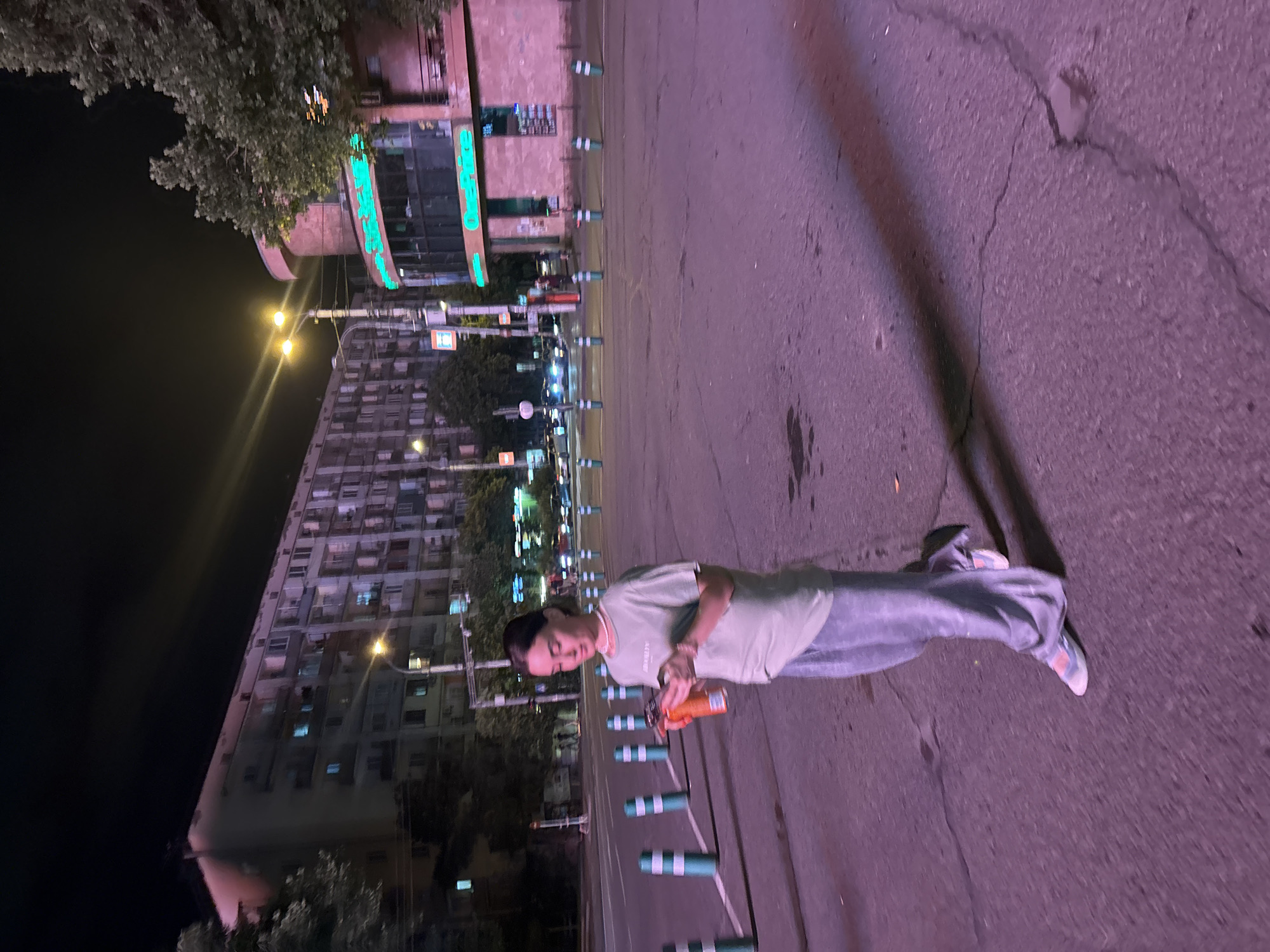 In Tbilisi, Georgia, at night, a young woman walks across a street. She is wearing a light-colored t-shirt and light-colored wide-leg pants. She is holding an orange cup in her left hand and looking at her phone in her right hand. The street is paved and has several rows of green and white bollards. In the background, there is a large, multi-story apartment building and a building with neon green signage. Streetlights illuminate the scene with a purple-pink hue. The overall impression is of an urban night scene in Tbilisi.