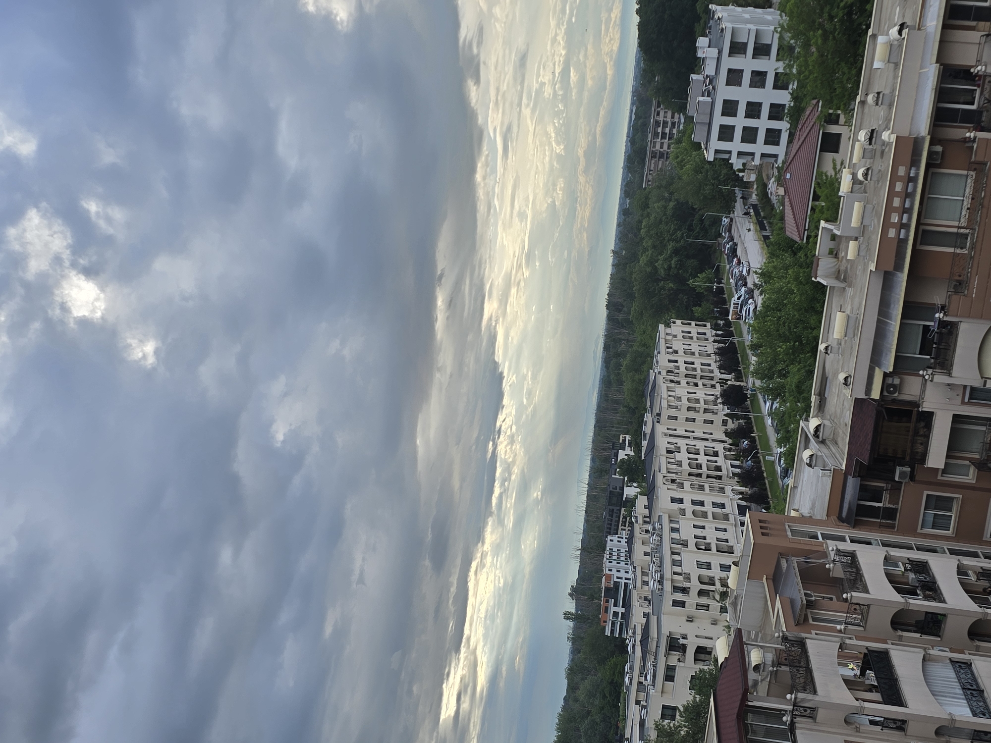 The image captures a high-angle view of a cityscape under a sky dominated by storm clouds. A lighter, brighter section of sky appears on the horizon, contrasting with the dark, heavy clouds above.
Below the sky, the urban landscape features a mix of buildings and green spaces. Several multi-story buildings are visible, exhibiting various architectural styles and colors. Some have flat roofs, while others have pitched roofs. Many windows and balconies are visible on the buildings.
Trees and vegetation are interspersed throughout the urban area, providing a touch of greenery among the structures. A road with parked cars runs through the landscape.
No people are directly visible in the image, focusing the scene on the interplay of natural and built environments.