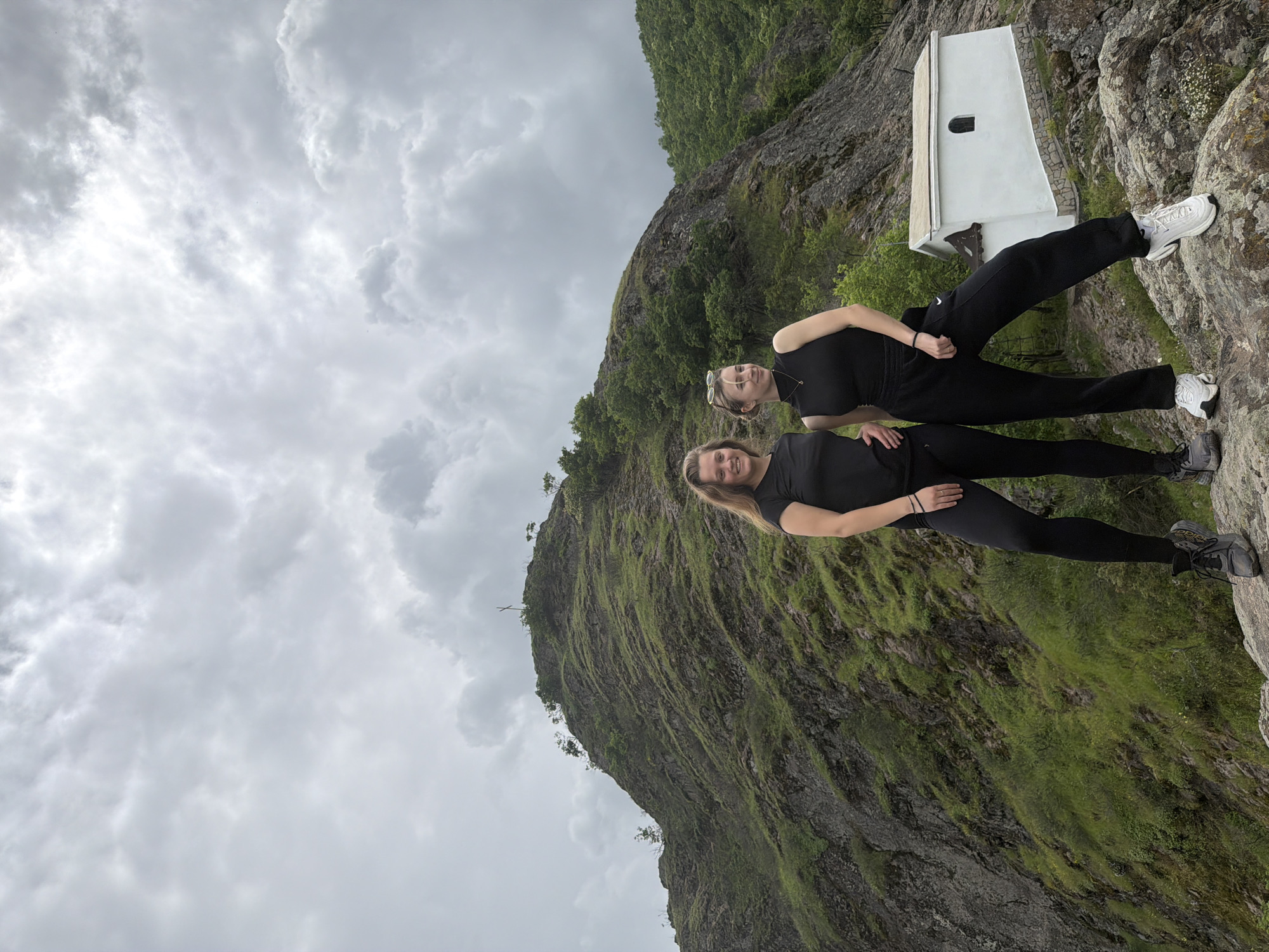 Two young women in black athletic wear stand on a rocky outcropping against a cloudy sky. They are positioned in the foreground of a steep, green mountainside. In the background, partially obscured by the mountain, is a small, white, rectangular structure with a dark doorway. This scene appears to be set in or near Ustina, Bulgaria, based on the surrounding landscape and architecture.