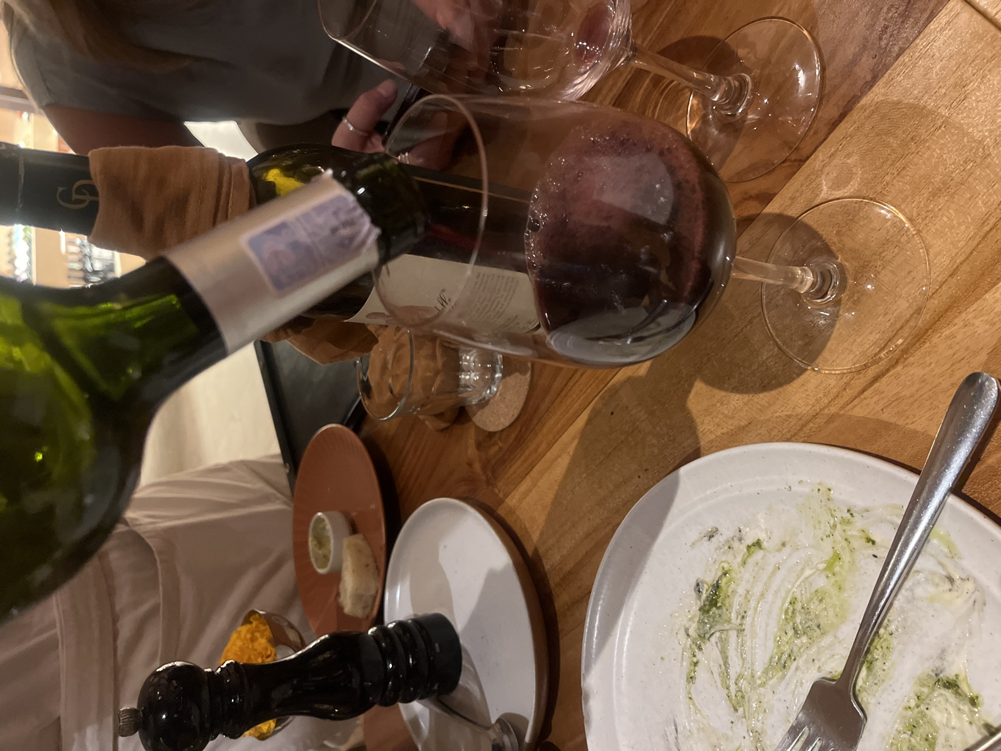 A bottle of red wine is being poured into a wine glass. Two glasses of wine sit on a wooden table. There are plates with remnants of food. A silver fork rests on one of the plates. There is a pepper grinder and a small dish with bread. The location is Kuta Selatan, Indonesia. There are two people in the scene.
