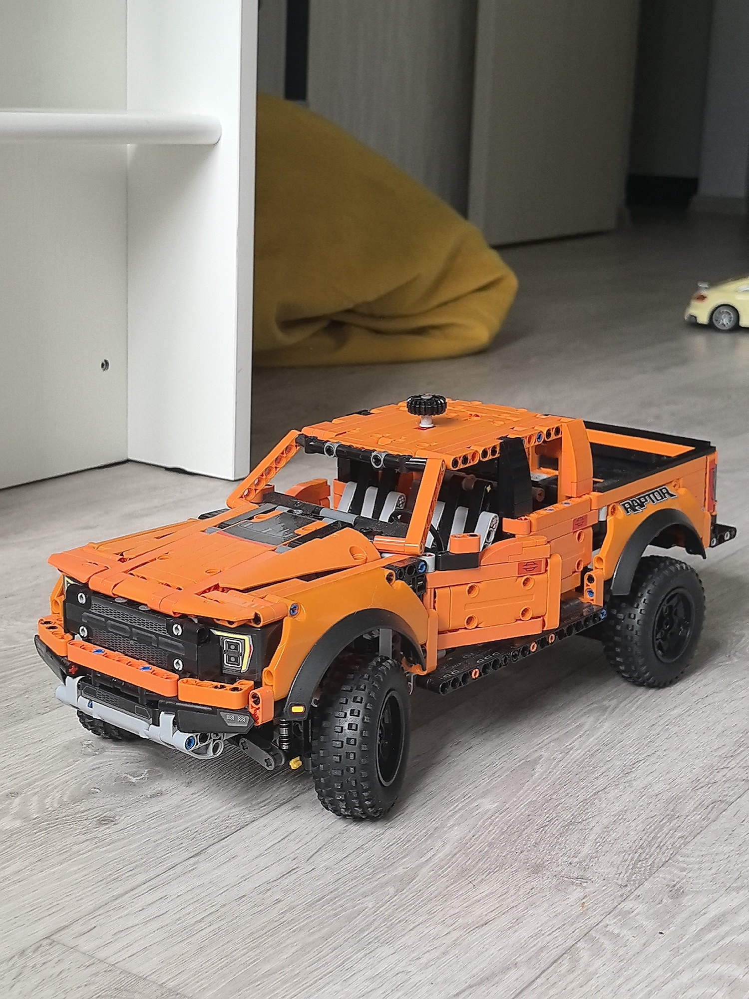 A low-angle, medium-shot depicts an orange Lego Technic Ford F-150 Raptor pickup truck positioned on a gray laminate floor. The truck is detailed with visible suspension, tires, cab, and "RAPTOR" lettering on the rear quarter panels.
In the background, out of focus, are a white shelf unit to the left, a yellow beanbag chair, and a small yellow toy car to the right.
The scene is likely indoors, based on the flooring and surrounding furniture. There is no indication of this being in Perm, Russia.