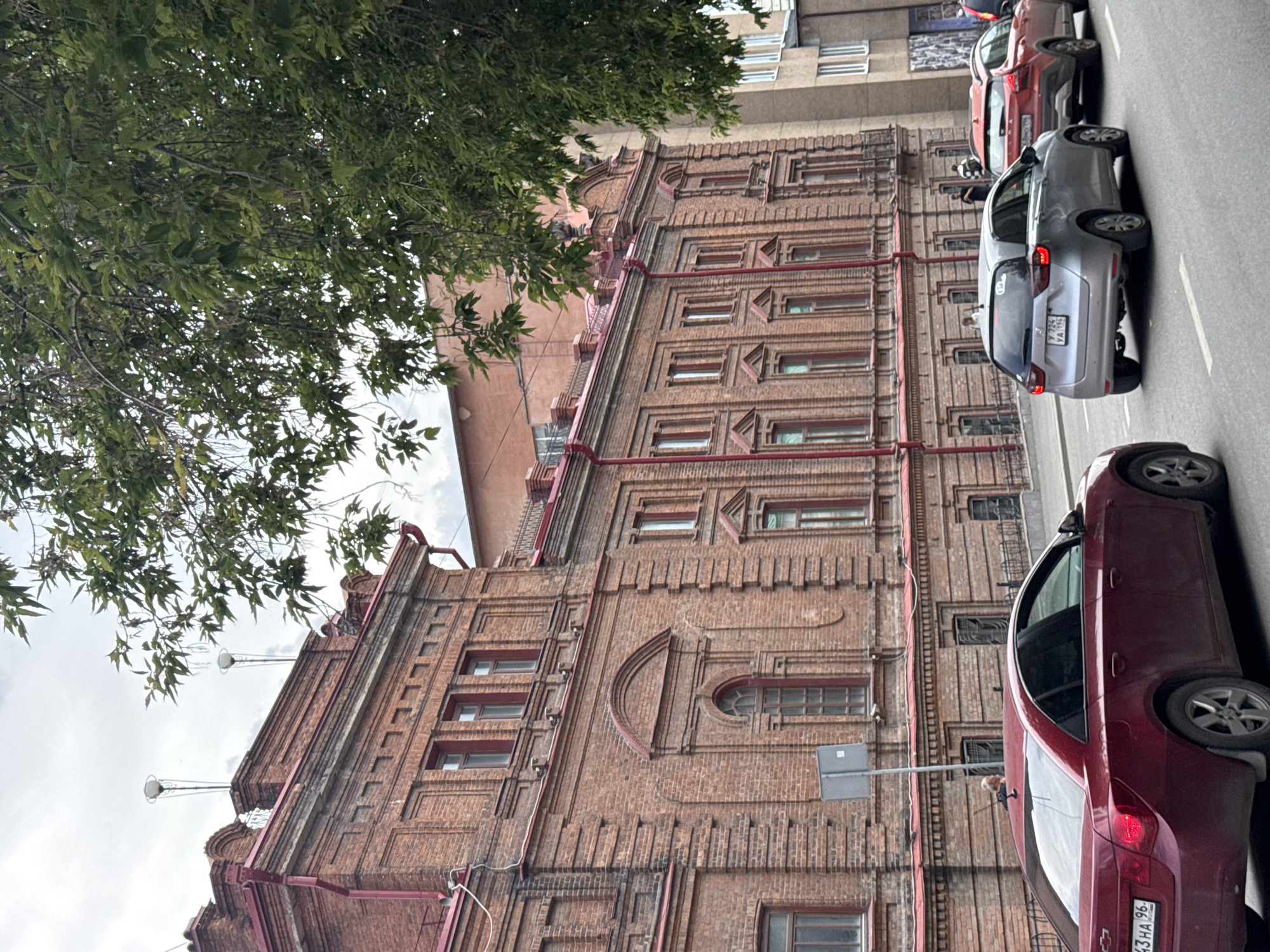 This media file captures a street scene in Yekaterinburg, Russia, featuring an old brick building with ornate architectural details. The building, constructed from reddish-brown bricks, has numerous windows, some with decorative arched frames. Red piping runs along the exterior of the building, and there are some modern additions like streetlights protruding from the roof.
A line of cars is parked along the street in front of the building. These include a dark red sedan in the foreground, a silver hatchback, and a red SUV further down the road, all facing away from the camera. The license plates of the cars are visible, with one reading "Y 724 YA 196".
A person can be seen standing on the roof of the dark red sedan, and another person is visible in the silver hatchback.
The scene is outdoors, under a cloudy sky, suggesting it might be overcast or a slightly dull day. The presence of a tree with green leaves in the upper left corner indicates it is likely spring or summer. The image is taken from a slightly elevated perspective, looking down the street.
The overall impression is a slice of urban life in Yekaterinburg, showing the interplay of historical architecture and modern vehicles. There is no specific event depicted, but rather a snapshot of a typical day.