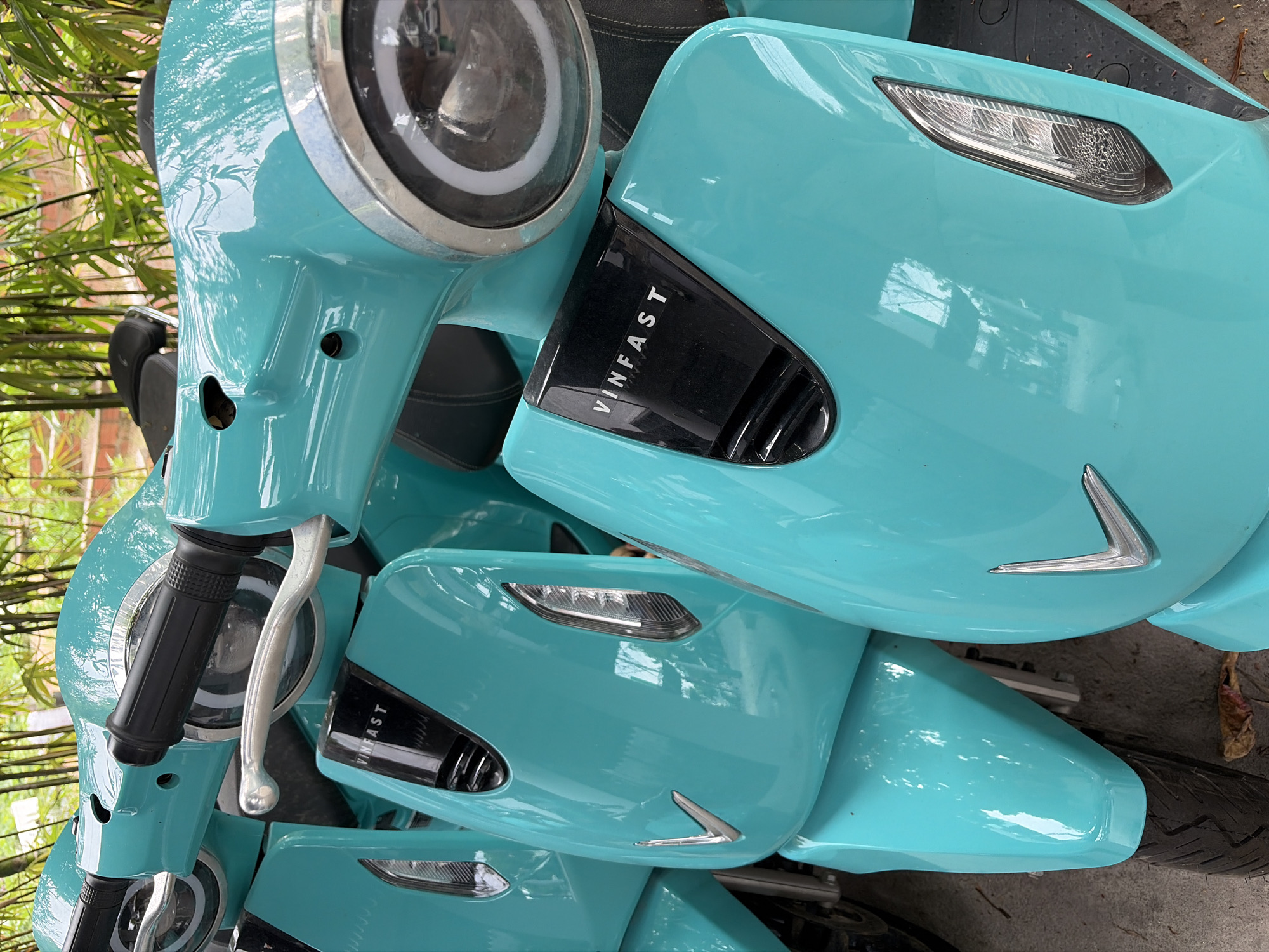 The image shows a close-up view of several turquoise electric scooters parked outdoors. The scooters are lined up, with the front of one scooter prominently featured in the foreground, angled slightly to the right. This scooter has a glossy turquoise body, a black "VINFAST" emblem on its front panel, and a silver V-shaped emblem below that. A silver brake lever and a black handlebar grip are visible on the left side. The scooter also features a clear headlight with a white ring of light. Behind the foreground scooter, parts of other identical scooters are visible, also in turquoise. The background consists of blurred green foliage, possibly bamboo, suggesting an outdoor setting with natural elements. The ground appears to be concrete or pavement, with some fallen leaves or debris visible. The lighting suggests it is daytime. The scooters appear to be part of a rental or shared mobility service, common in cities like Đà Nẵng, Vietnam.