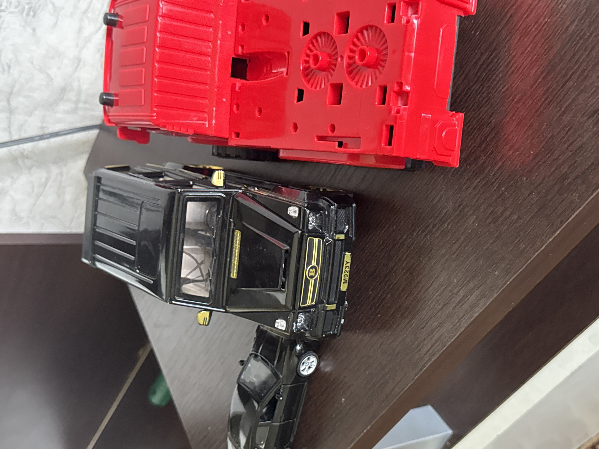 This is a photograph of two toy vehicles. The top subject is a red toy truck. The bottom subject is a black toy car, possibly a limousine or a van, and it is partially obscuring a smaller, darker toy car. The toys are placed on a dark brown, wooden surface. The background is out of focus and appears to be a light-colored textured surface on the left and a lighter surface on the right. There is a visible label on the back of the black toy car that reads "M923Y". The image appears to be taken indoors, with no indication of time of day or weather. The setting is likely a home or a display area for toys. No people or specific emotions are discernible.