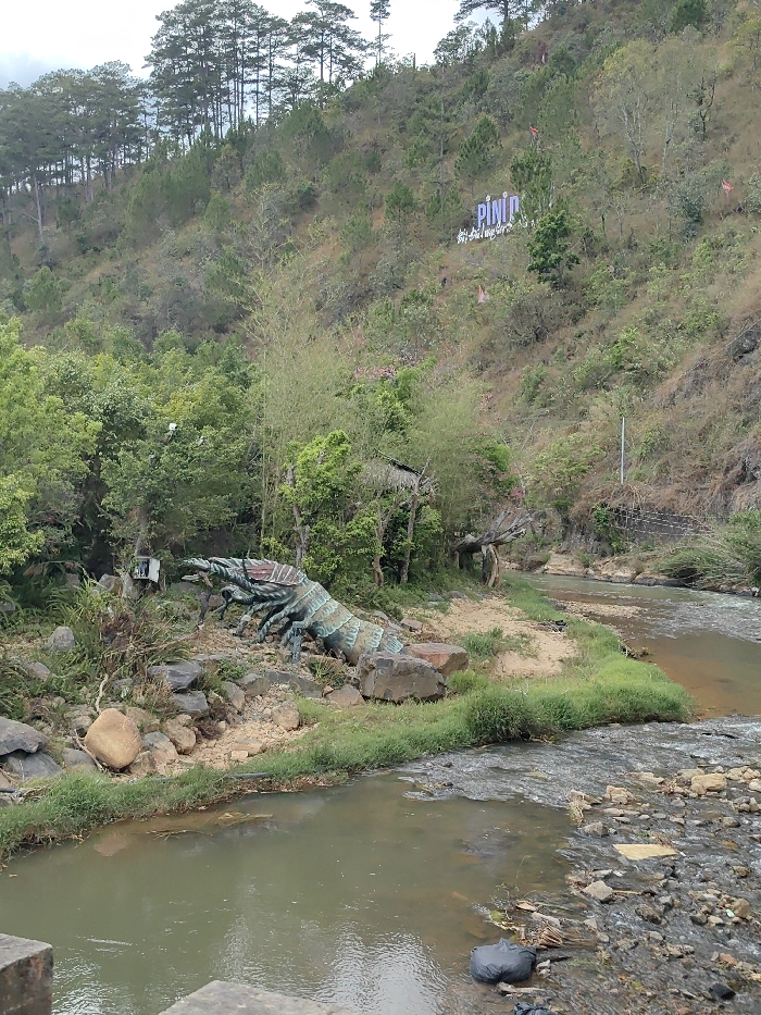 The image captures a diverse outdoor landscape featuring a river, a sculptural element, and a steep, tree-covered hillside.
In the foreground, a river flows, segmented into two main parts. On the left, the water appears calmer and reflects the light, while on the right, it is shallower, rockier, and shows more movement. A narrow, grassy bank separates these two sections of the river. Scattered rocks and pebbles line the banks, and some debris, including a black plastic bag, is visible in the water on the bottom right. Part of a dark, sturdy structure, possibly a bridge or viewing platform, is visible in the very bottom left corner.
Dominating the midground on the left bank of the river is a large, multi-segmented sculpture resembling a giant crustacean or insect. It is predominantly greenish-blue with visible darker bands and lighter accents, appearing to be made of metal or a similarly durable material. The sculpture features multiple legs and antennae and is positioned on a sandy, rocky patch of ground amidst natural vegetation. Behind the sculpture and extending up the bank, there is a dense growth of green trees, bushes, and some tall, thin bamboo-like stalks. Partially obscured by the foliage behind the sculpture is a rustic structure with a natural or thatched roof, possibly a small hut or shelter. To the left of the sculpture, a white, box-like object, possibly an old appliance or container, is visible.
The background is dominated by a steep hillside rising above the river. The lower slopes are covered in a mix of sparse brown and green vegetation, suggesting a somewhat dry environment or season. Further up, the hillside becomes densely forested with numerous tall, slender conifer trees, likely pines, which crest the hill against a bright, overcast sky. Prominently displayed on the upper part of the hillside, amidst the sparse vegetation, is a large, light-colored sign that clearly spells out "PININ." Below this main text, smaller, less legible text appears to read "Happy and Love..." followed by an unclear word. Several small, red or orange flag-like decorations are also scattered on the hillside near the sign.
The scene appears to be set during daytime, under a bright, possibly overcast sky. There is no explicit activity taking place, suggesting a static view of a natural or landscaped park area that incorporates both natural elements and artificial attractions.