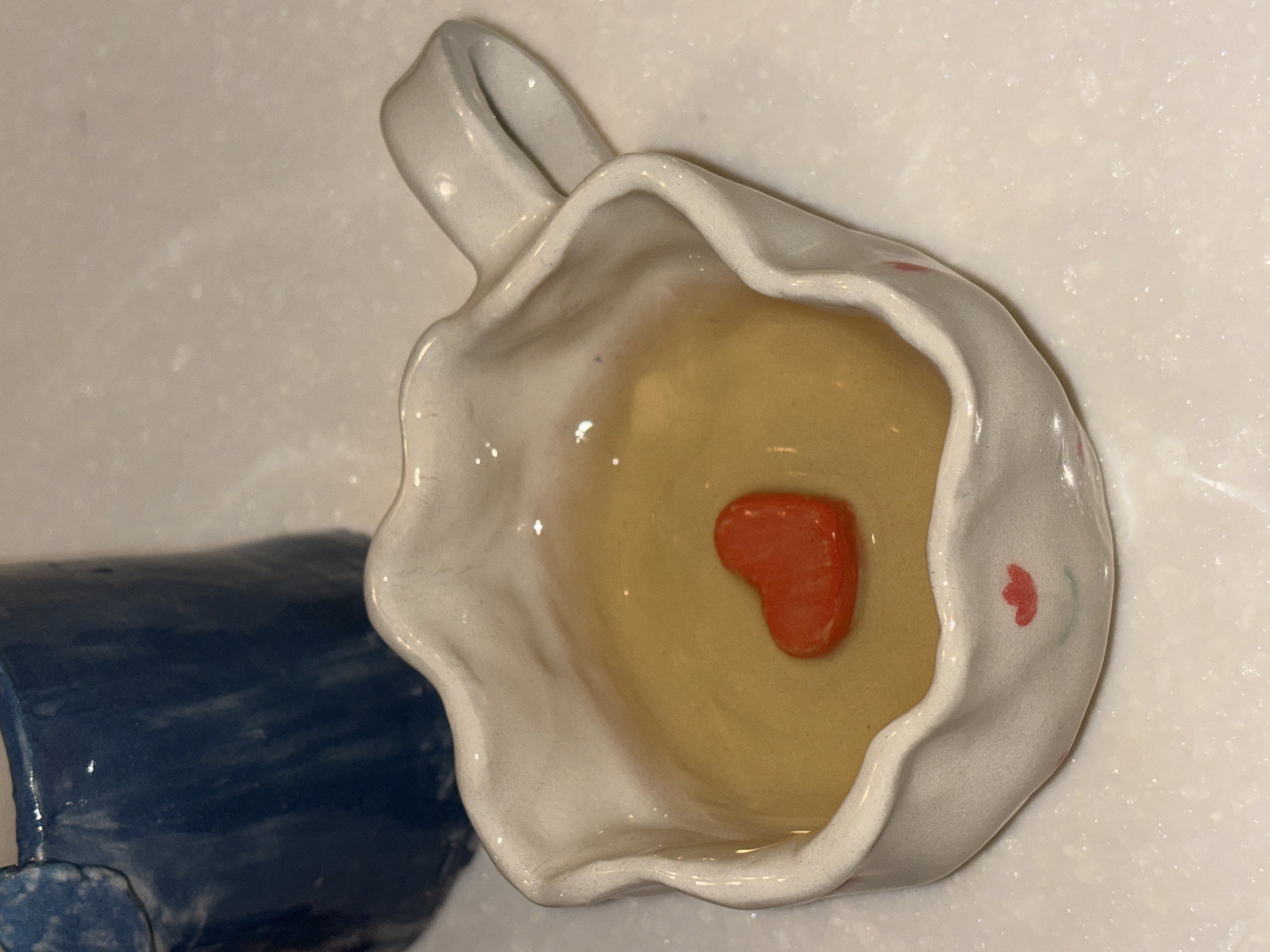 A high-angle, indoor shot depicts a white, ceramic, handled cup lying on its side against a blue, cylindrical object, against a white surface. The cup is irregularly shaped with a ruffled rim. Inside the cup is a yellow liquid with a single, red heart-shaped object resting at the bottom. A small, red floral design is painted on the side of the cup. The scene is located in Choeng Thale, Thailand.