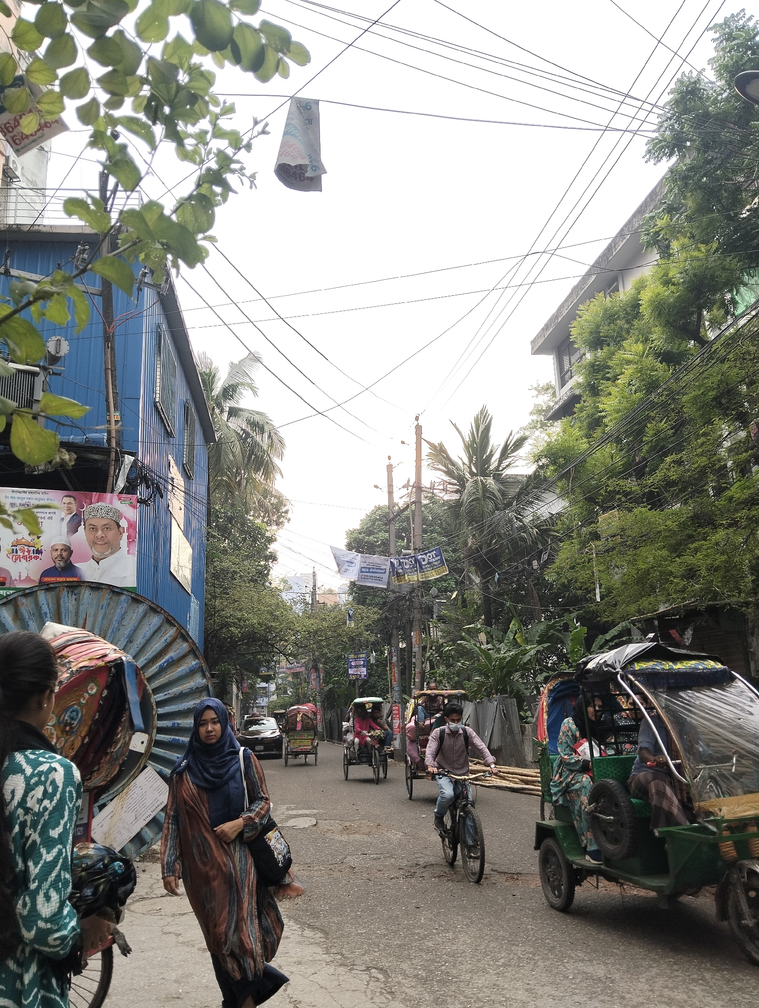 The image shows a street scene in Dhaka, Bangladesh. A woman in a blue hijab and patterned dress walks towards the camera on the left sidewalk. A woman with long black hair is next to her, also with a patterned dress. A man wearing a mask is riding a bicycle toward the camera. Several rickshaws and cars are visible on the street, with people riding in some of the rickshaws. Buildings and trees line the sides of the street, with power lines crisscrossing overhead. There are campaign posters on some buildings and utility poles. Some posters have the name "DODOT".