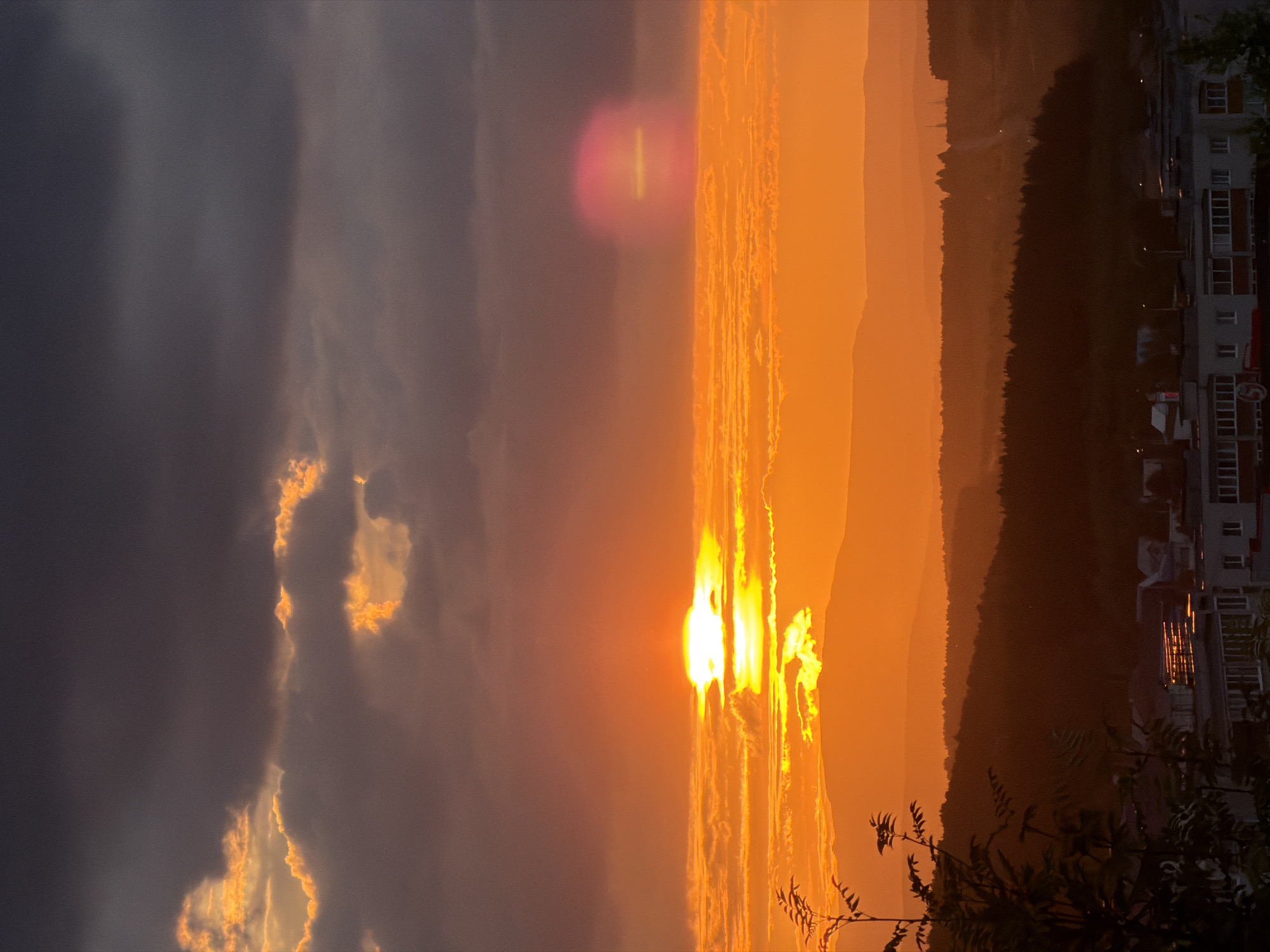 A vertical, wide-angle, close-up shot captures a dramatic sunset over a city. The sun, a bright white orb with a yellow corona, is partially obscured by low-lying clouds tinged with orange and yellow. Dark, ominous clouds fill the upper portion of the frame, creating a stark contrast with the vibrant sky. A pinkish lens flare hovers to the right of the sun. In the foreground, silhouetted trees with sparse foliage frame the left side of the image. Below the horizon, layers of receding hills and a dark forest are visible. The lower third of the image shows a residential area with buildings, some featuring balconies, bathed in the warm, orange light of the setting sun. A few distant lights and a road winding through the landscape can be seen. The overall impression is one of a breathtaking natural phenomenon occurring above a populated area.