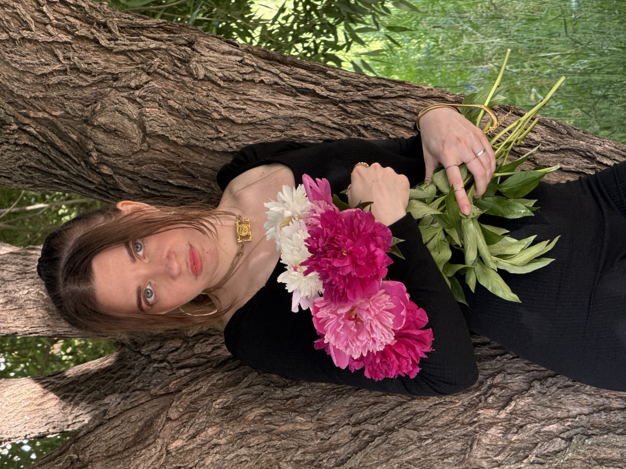 A woman is lying on her back on a large tree trunk, her head resting on the bark. She is wearing a black long-sleeved top and has a bouquet of pink and white peonies in her hands. A gold necklace with a rectangular pendant is visible around her neck, and she is wearing several rings on her fingers and a gold bracelet on her wrist. Her hair is brown, and she is looking up at the camera with blue eyes and pink lips. The background is blurred green foliage, suggesting an outdoor setting, possibly a park or forest. The lighting suggests it is daytime, with dappled sunlight filtering through the leaves. The woman's expression appears calm and serene. There is no visible text in the image.