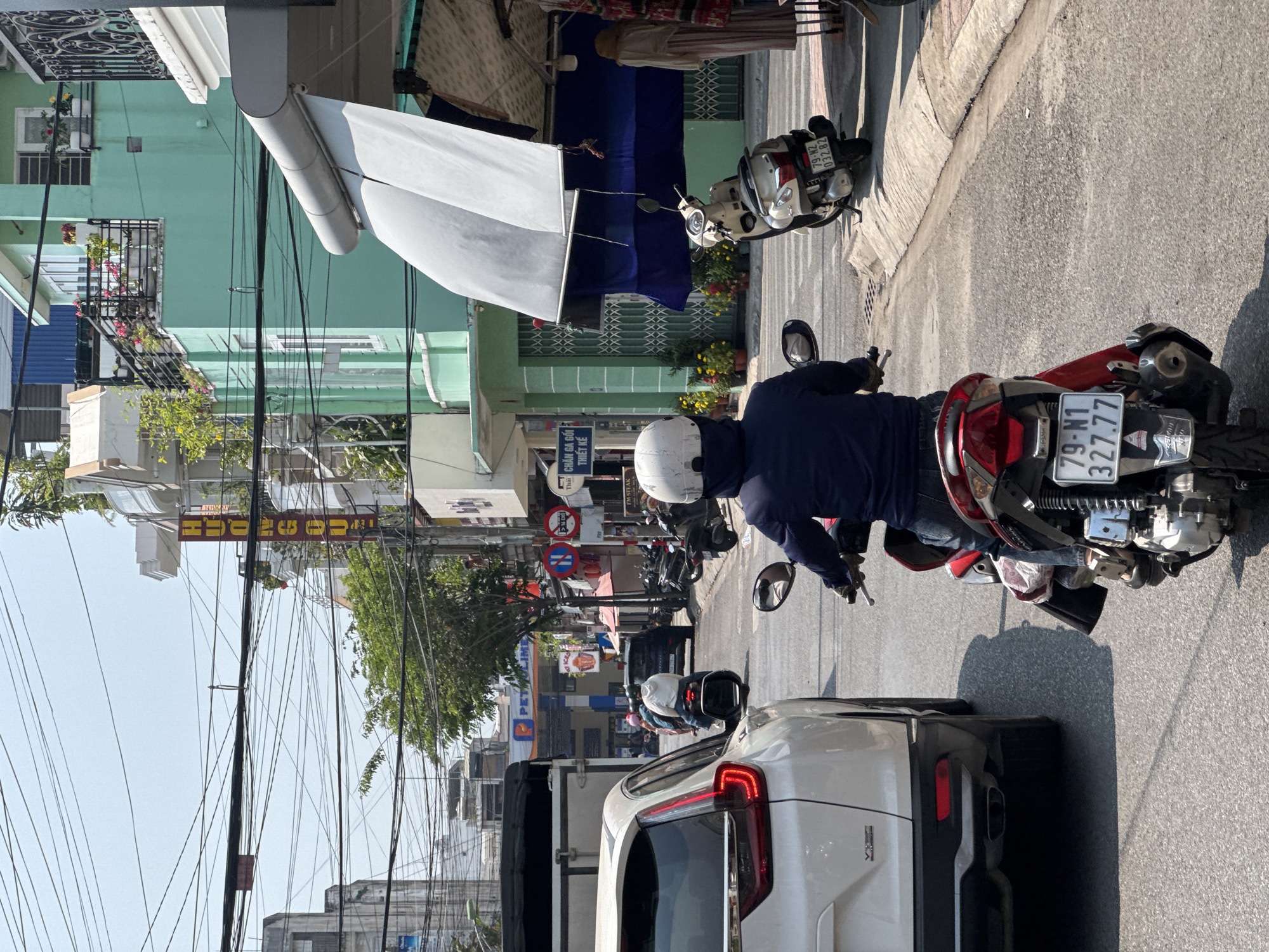 The image captures a street scene in an urban environment. A person on a red motorcycle, wearing a white helmet and blue jacket, is in the foreground. The motorcycle's license plate reads "79-N1 327.77."
In front of the motorcycle is the rear of a white SUV. The background shows a street lined with buildings. A green building with flowers is visible on the left, along with an awning. Various power lines stretch across the sky. Street signs and a sign for "PET" are seen in the distance. Another motorcycle is parked along the street, with a license plate reading "79-N2 032.82." A sign for "I'M STEAK" and "CHĂN GA GỐI THIẾT KẾ" is also visible.