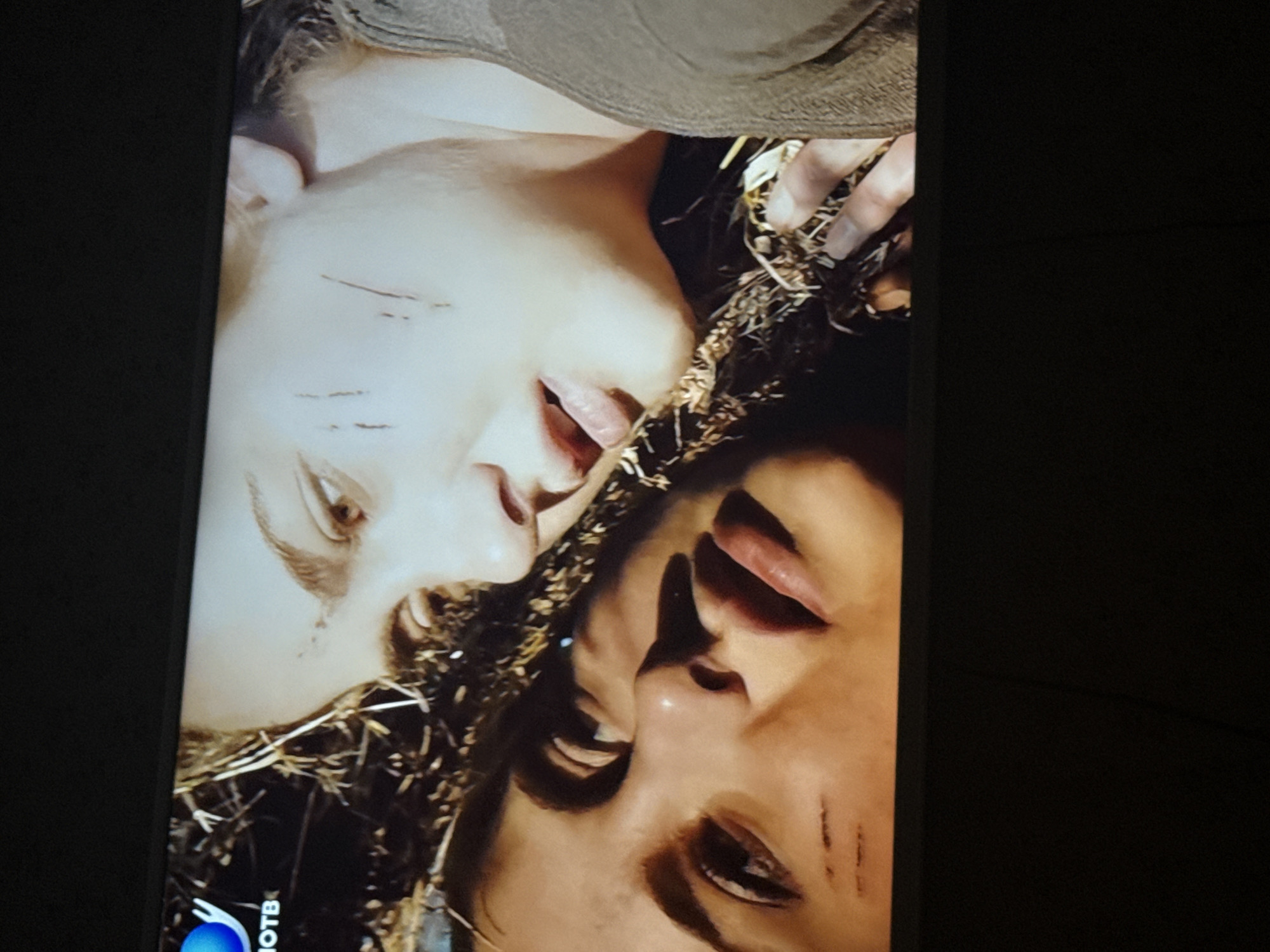 The image shows two people lying close together in what appears to be straw or dried grass. Both individuals have fair skin and marks resembling cuts on their faces. The person on the left has lighter hair, and the person on the right has darker hair. Their faces are oriented towards each other. The background beyond the straw is dark. The location is not discernible from this image alone, so there is no information pertaining to the city of Mytishchi, Russia.
