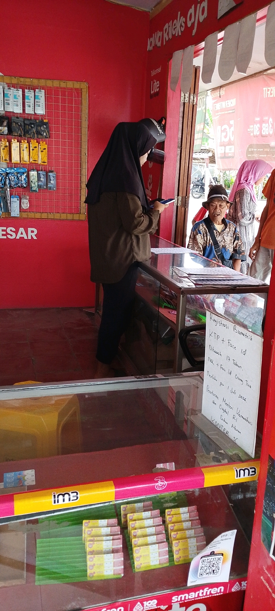 The image depicts a small roadside stall, likely a mobile phone shop, in Kertahayu, Indonesia. A woman wearing a black hijab and an olive-green jacket stands at the counter, looking at her phone. She is positioned to the left of the frame. In the background, an elderly man with a patterned shirt and a dark hat is seated, looking forward. Behind him, another person in a pink hijab is partially visible.
The stall's interior is red, with shelves displaying various mobile accessories like earphones. The counter is made of glass, revealing rows of SIM card packages and some green banana leaves. A white sign is placed on the counter, listing regulations for biometric registration, including "KTP + Face ID," "Dibawah 17 Tahun," and "NIK + Face Id Orang Tua." The sign also mentions "Peraturan Menteri Komunikasi dan Digital RI" and "KONTER" which likely refers to the stall.
Outside the stall, through an open doorway, a motorcycle is parked. The overall scene suggests a typical day at a local business in a residential area. The lighting indicates it's daytime.