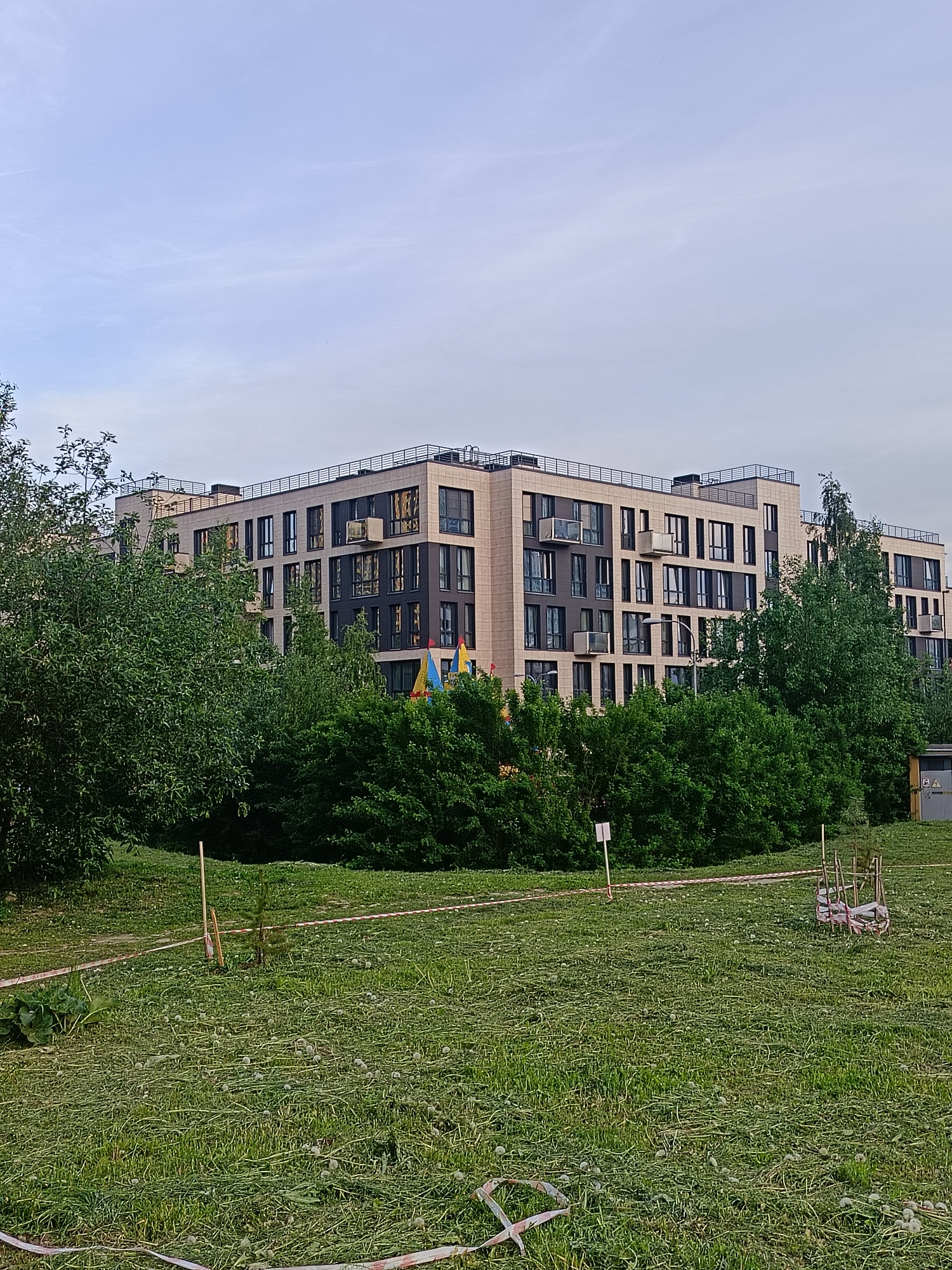This image captures an urban residential scene in Saint Petersburg, Russia, under a pale blue sky with light, scattered cloud cover. In the background, a modern, multi-story building constructed with light beige masonry cladding and dark grey window panels is prominent. The building features numerous vertical windows, some incorporating small balconies with dark metal railings, and several external air conditioning units are mounted on its facades. The roofline is equipped with a dark protective railing. Partially visible behind the trees on the left are colorful elements, indicative of playground equipment. The midground is characterized by a dense, verdant expanse of various deciduous trees and bushes, obscuring the lower levels of the building. The foreground comprises a recently mowed lawn, covered extensively with fresh grass clippings and numerous white dandelion seed heads. A narrow, unpaved pedestrian path traverses the grassy area. Within the foreground, a young sapling is anchored by a wooden stake, and several additional wooden stakes are sparsely distributed. Sections of red and white striped barrier tape are lying loosely on the mowed grass. To the right, a small, open-sided utility cabinet, primarily yellow and grey with a warning symbol, is situated at the edge of the lawn. Adjacent to this, a small, rough wooden frame or structure is present. No human or animal subjects are discernible within the frame.