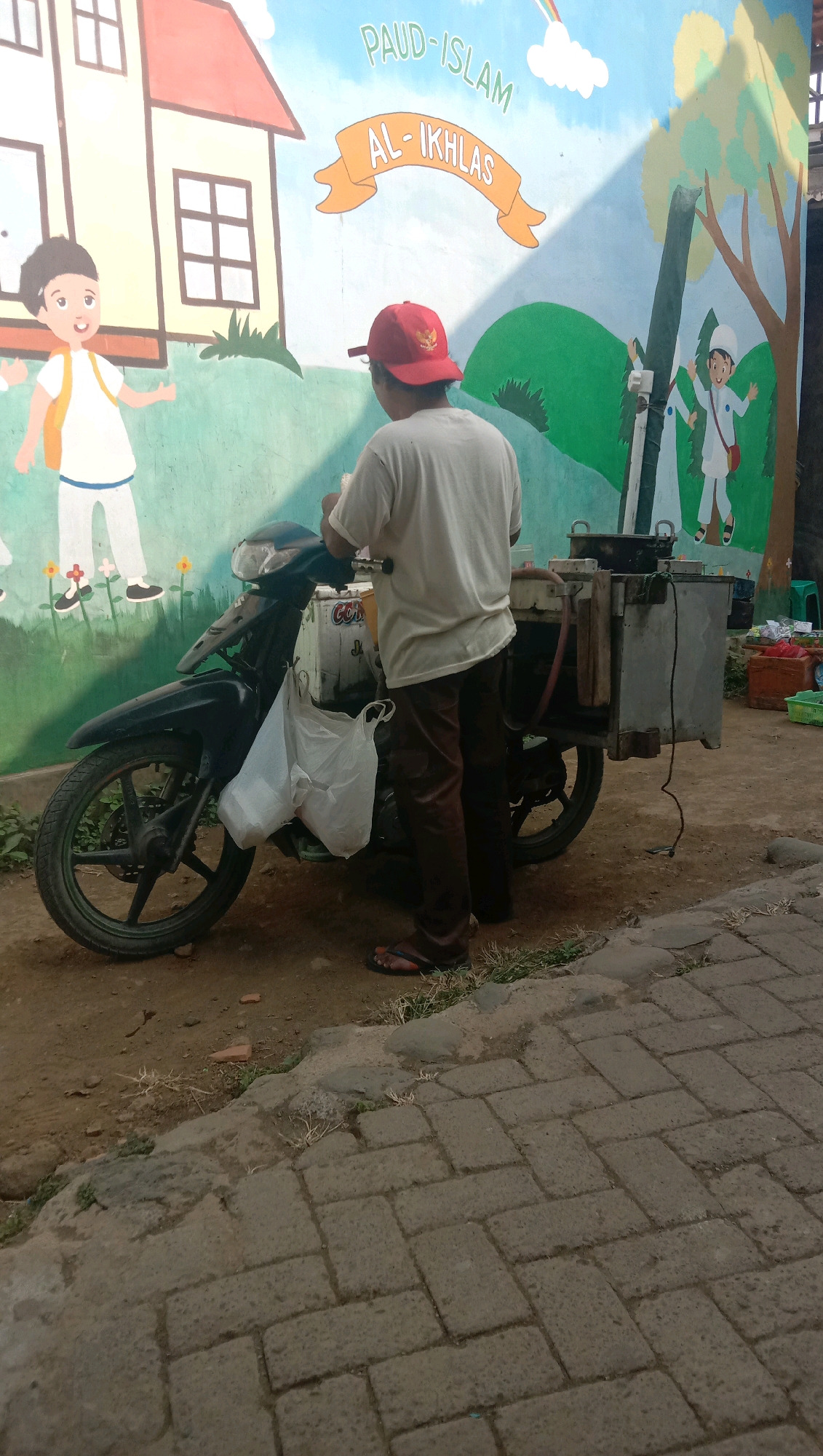 A person wearing a white t-shirt and brown pants, with a red cap, stands next to a black motorcycle with a food cart attached to the back. The food cart is made of metal and contains various containers and utensils. A white plastic bag is hanging from the motorcycle's handlebar. The person appears to be preparing or serving food from the cart. The background features a colorful mural painted on a wall, depicting children, buildings, trees, and the text "PAUD-ISLAM AL-IKHLAS". The ground is a mixture of dirt and brick paving stones. The scene appears to be outdoors, possibly in front of a school or public area. The location is identified as Karang Anyar, Indonesia.