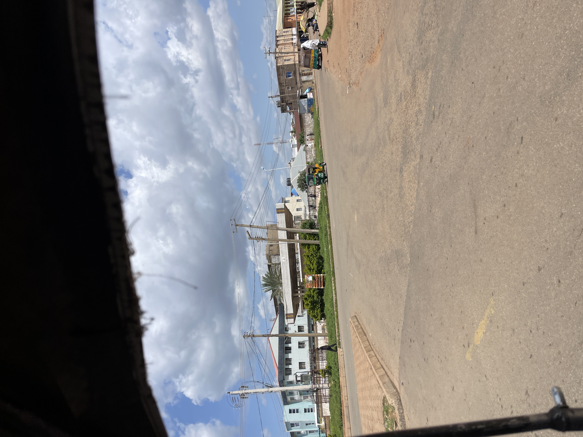 The image depicts a street scene in Rafin Par, Nigeria, on a partly cloudy day. The perspective suggests the photograph was taken from inside a vehicle, likely a tuk-tuk, as evidenced by the dark frame of the vehicle's structure in the foreground.
The road is paved with asphalt and appears to be a junction. There are buildings lining the street on both sides. On the left, a light blue multi-story building with multiple windows stands prominently, accompanied by utility poles and overhead wires. A person in dark clothing is walking on the sidewalk in front of this building. Further down the street on the left, there are more buildings, some appearing to be residential or commercial, with white awnings and fenced areas.
On the right side of the road, there are more buildings of varying architectural styles and heights. One building has a beige facade with arched windows, and another appears to be a newer structure with a grey facade. Several people are visible on the right side of the road, near a yellow umbrella.
Two tuk-tuks, common modes of transportation in Nigeria, are present in the scene. One is green and white and is moving on the road. The other, also green and white, is parked or moving slowly on the right side of the road, near the people under the umbrella.
The environment includes green vegetation, primarily bushes and some trees, planted along the sides of the road and in front of some buildings. The sky is a mixture of blue sky and white cumulus clouds, indicating dynamic weather conditions. The overall impression is a bustling urban environment with typical infrastructure and daily life activities.