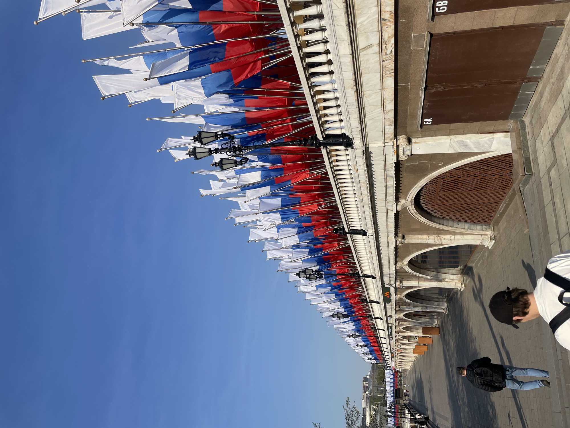 The image shows a building facade in Moscow, Russia, adorned with numerous Russian flags. The building has a row of arched openings at street level, with the flags positioned along the upper edge of the facade. White decorative elements form a balustrade along the roofline. The sky is clear blue. Two people are visible in the foreground, one standing and another walking in the opposite direction, suggesting a street scene. The numbers "6A" and "6B" are visible on what appear to be metal doors in the facade. Based on the architectural style and the presence of Russian flags, the building is likely located in a central area of Moscow, possibly near Red Square.