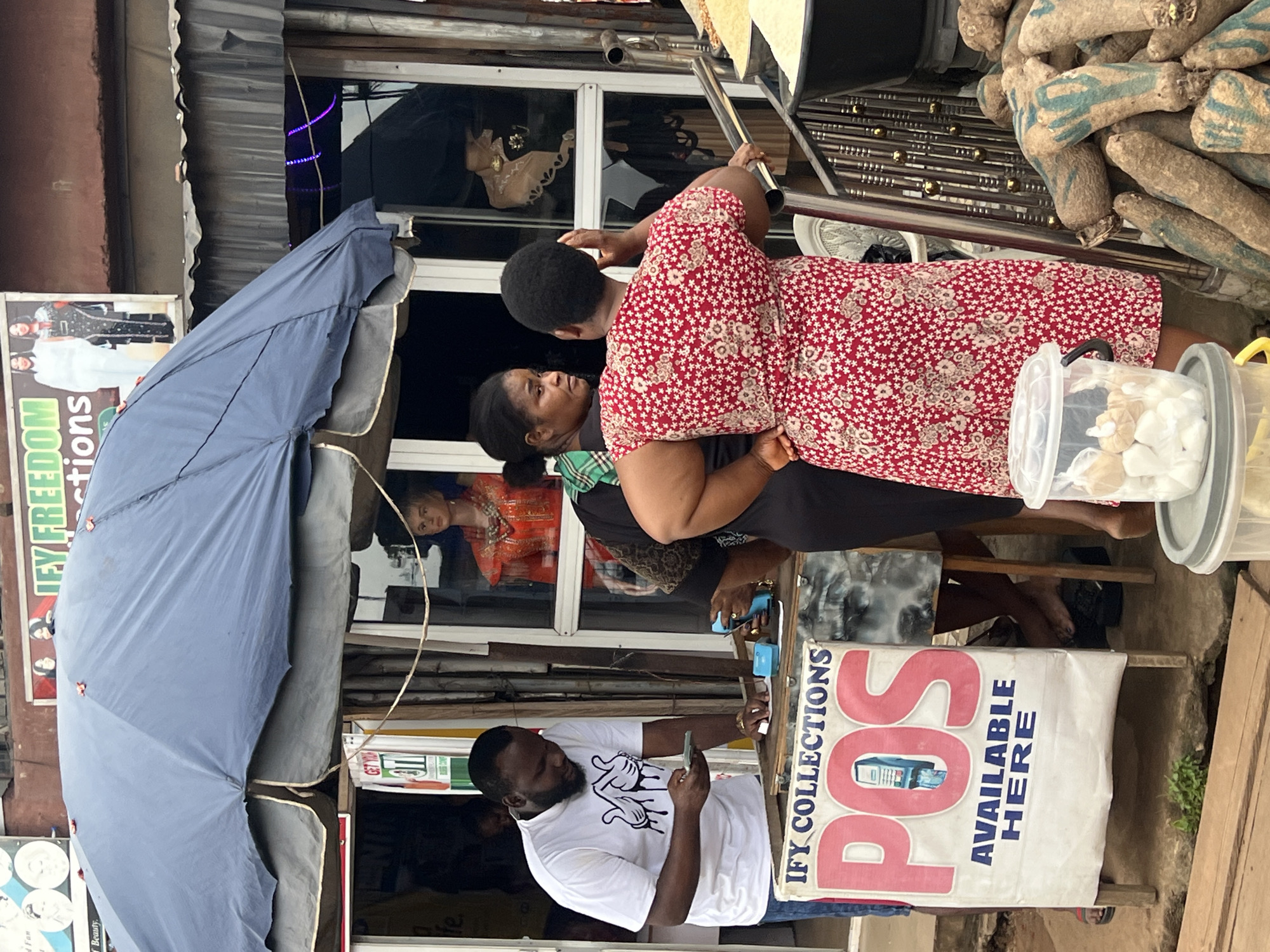 The image depicts an outdoor scene in Nkpor, Nigeria, likely a street-side market or vendor stall. The primary subjects are a group of people interacting, with a focus on two women and a man. One woman, wearing a red and white floral dress, is facing away from the camera, her back towards a window. Another woman, in a green and black checkered top, is facing the first woman, her head turned to the side. A man wearing a white t-shirt with a cartoon graphic is also visible, looking down at his phone.
The setting is a busy street, with a building facade featuring a window where other individuals are faintly visible. There's a large blue umbrella on the left side of the frame, partially obscuring the view. Objects like a metal railing, a plastic container with white items (possibly food or merchandise), and a white banner are also present. The banner displays text that reads "IFY COLLECTIONS POS AVAILABLE HERE" and features an image of a point-of-sale machine. Another poster to the left of the umbrella shows two women in traditional attire.
The image appears to be taken during the daytime, with natural lighting. The presence of a market suggests a commercial activity is taking place, likely the exchange of goods or services. The people's postures and engagement with their phones suggest casual interaction and daily life activities. The overall atmosphere seems lively and indicative of a bustling Nigerian marketplace.