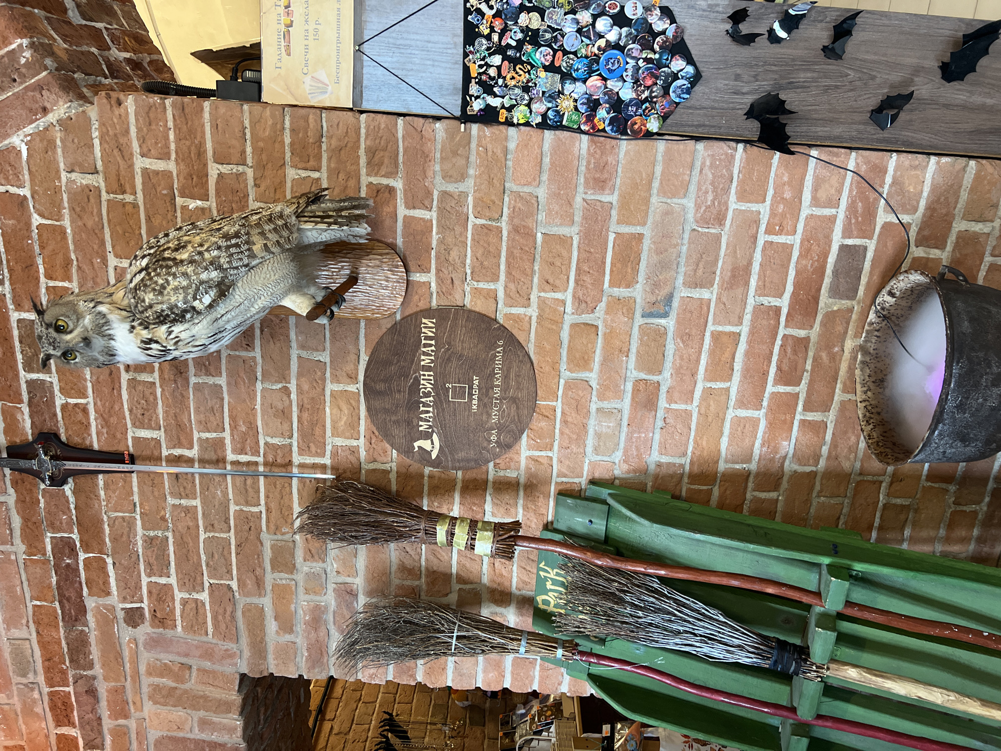 This media file captures an indoor display with a clear magic or fantasy theme, set against a rustic red brick wall. The scene is brightly lit, with no indication of time of day or weather as it's an interior shot.
Prominently featured on the left is a taxidermied owl, meticulously preserved with detailed plumage, perched on a small wooden mount. Below the owl, a decorative sword with an intricately designed hilt is horizontally mounted on the brick.
In the center, a circular wooden sign is affixed to the wall, bearing Russian text: "МАГАЗИН МАГИИ" (Magic Shop), "КВАДРАТ," and "УФА МУСТАЯ КАРИМА 6," providing the name and address of the establishment in Ufa, Russia.
To the right, two traditional-looking brooms with natural twig bristles and dark wooden handles are neatly arranged on a green wooden rack. In the upper right corner, a large, dark cauldron emits a soft purple glow and mist, suggesting a magical brew or special effect. Above the cauldron and on a wooden shelf, several black bat cutouts are visible, along with a display board covered in numerous colorful pins or badges.
Further text is visible on a light-colored cardboard sign in the top left, advertising "Гадание на..." (Fortune-telling on...), "Свечи на жела..." (Candles for wish...), "150 р." (150 rubles), and "Беспроигрышная л..." (Win-win l...). The overall arrangement points to a retail store or themed attraction specializing in magical and fantasy items.