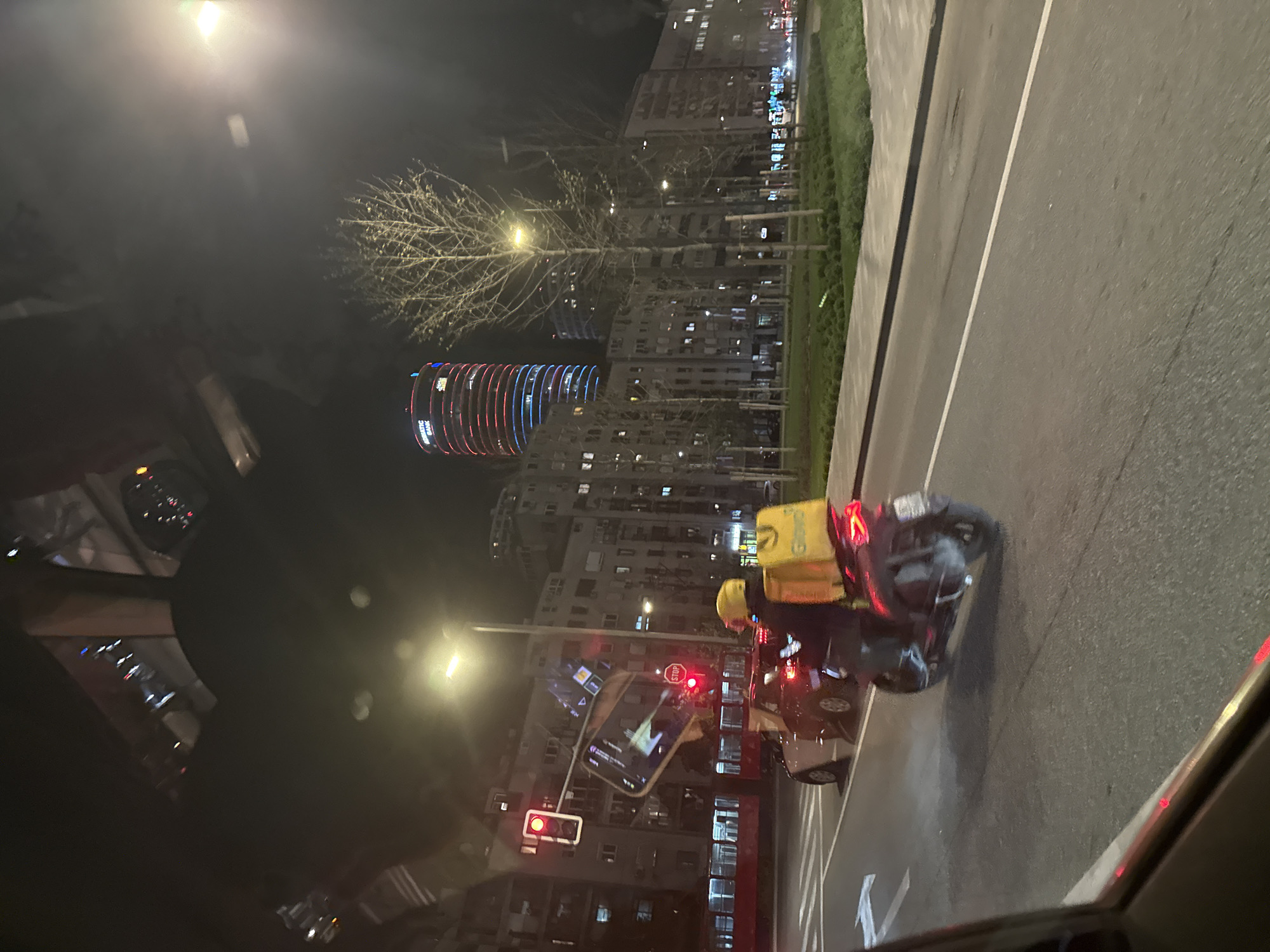 The image shows a night scene in Belgrade, Serbia, viewed from inside a car. A delivery person on a scooter with a yellow delivery backpack is stopped at a red traffic light. Cars are stopped behind the scooter. The skyline features residential buildings and a prominent, illuminated building with red and blue horizontal stripes. A smartphone is being held up in front of the traffic light. Trees are visible along the roadside. The street is wet.