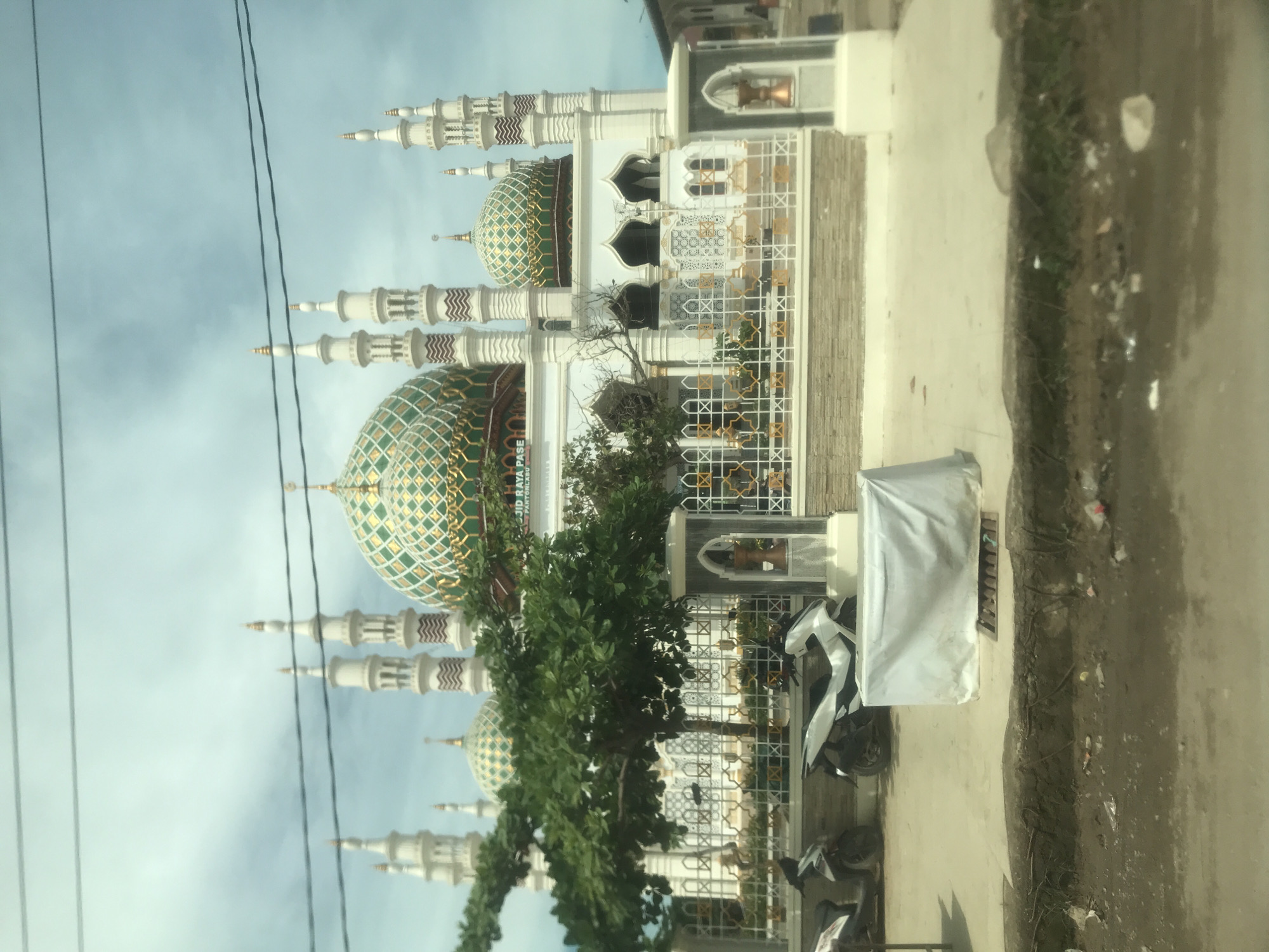 The vertically oriented image, captured during the daytime in Kotapantonlabu, Indonesia, presents a detailed view of a mosque. The mosque stands out with its pristine white structure, featuring multiple slender minarets topped with finials and adorned with distinct brown and white chevron patterns. Several large domes, beautifully tiled in an intricate green and gold diamond pattern, crown the mosque.
In the midground, a vibrant green tree partially screens the lower section of the mosque, while ornate white fencing with gold geometric designs borders the mosque's premises. Parked in front of the fence are several motorbikes, including one distinctively white and black vehicle, and another covered by a white tarp next to a white rectangular unit. Overhead, two dark electrical wires traverse the light blue, partly cloudy sky.
The foreground shows an unpaved strip of ground, littered with some small debris, running alongside a paved area. A visible banner or sign affixed to the mosque structure clearly displays the text "JAYA PASE" and "PANTUNLABU," providing a specific location cue. The overall scene is bright, devoid of people, and highlights the architectural beauty of the mosque against a clear sky.