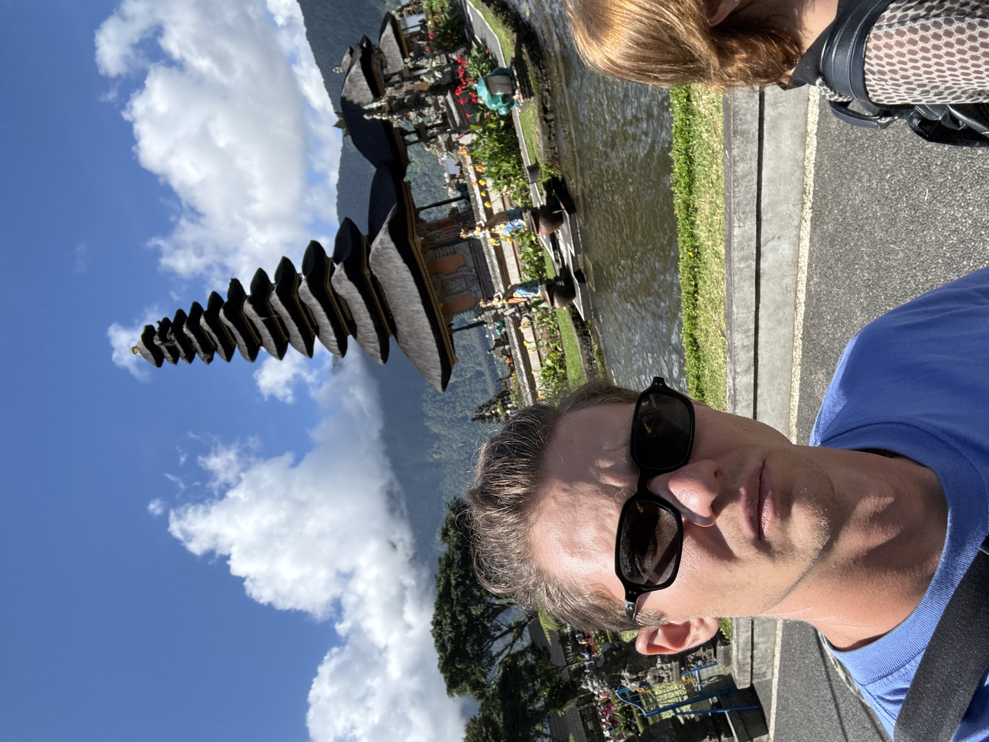 A selfie perspective captures a man in sunglasses and a blue shirt against the backdrop of Pura Ulun Danu Beratan, a Hindu temple in Baturiti, Indonesia. The temple, with its tiered pagoda-like structure, stands beside a body of water. A light-haired individual is partially visible in the upper-right corner. The sky is bright with scattered clouds.