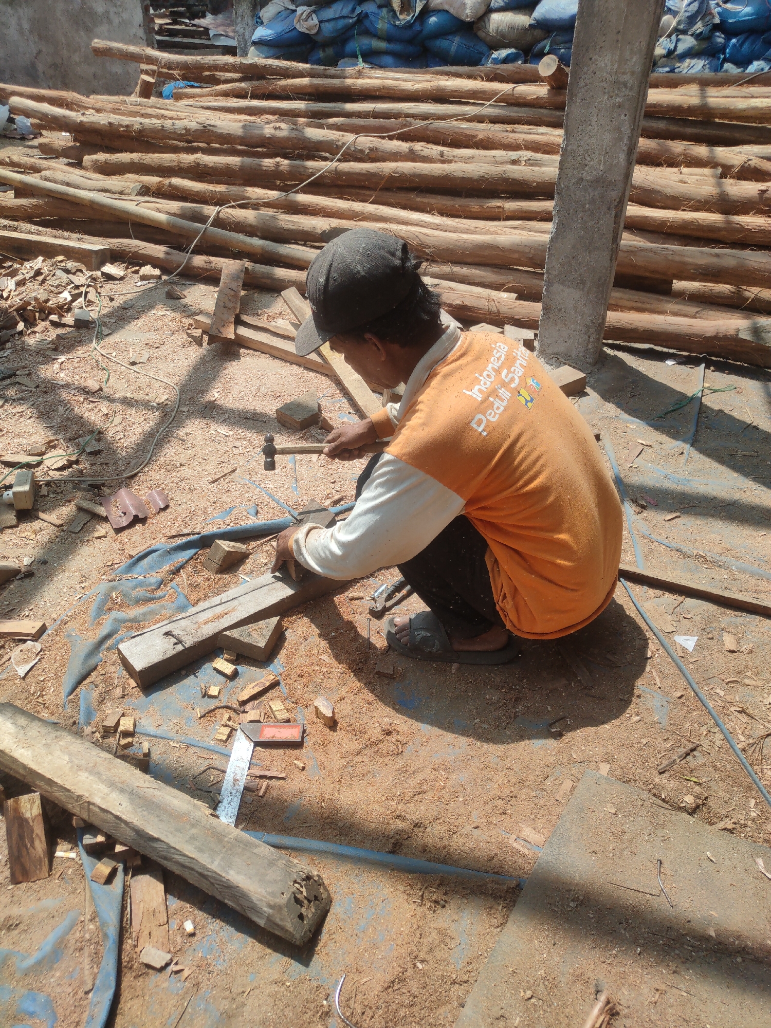 The image depicts a construction site in Sumedang, Indonesia, possibly a rural area or a developing part of the city. A person, wearing an orange t-shirt and a black cap, is squatting on the ground, actively engaged in carpentry work. They are holding a hammer in their right hand and appear to be hammering a nail into a wooden plank. The ground is covered with sawdust and wood scraps, indicating recent woodworking activity. In the background, a large pile of logs is stacked, suggesting a source of raw material for construction or woodworking. Blue tarpaulin sheets are visible under the logs and scattered around the work area, possibly to protect materials or provide a cleaner workspace. The scene is lit by bright sunlight, casting shadows on the ground. The overall environment suggests an outdoor, manual labor setting.
