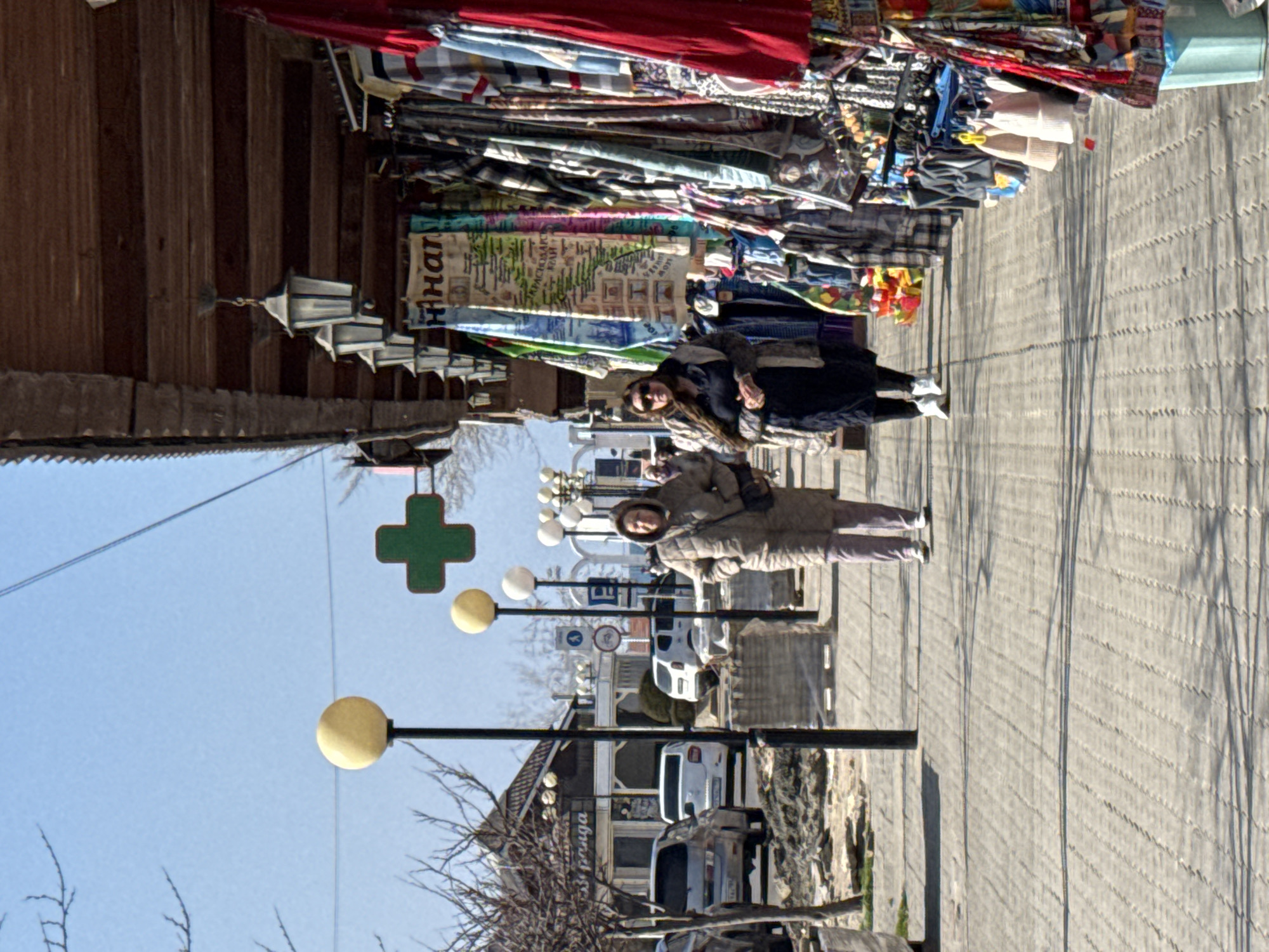 The image presents a daytime street scene in Anapa, Russia, identifiable by the Cyrillic text "АНАПА" visible on merchandise. Two adult females walk towards the viewer on a grey paved sidewalk, slightly to the right of the frame's center.
The female on the left wears a light brown quilted hooded jacket, dark trousers, and white athletic shoes, carrying a dark shoulder bag. The female on the right wears dark sunglasses, a dark top, a grey-brown jacket, and white athletic shoes. Both cast long shadows onto the sidewalk, indicative of a clear, sunny day.
To the right, a series of commercial stalls operate under a dark wooden overhang adorned with multiple antique-style hanging lanterns. Various textiles, clothing items, scarves, and hats in diverse colors and patterns are displayed on racks. A light blue bucket is visible at the lower right corner.
To the left, beyond the sidewalk, a street contains parked vehicles, including a silver SUV and a white sedan. Bare-branched deciduous trees line this side of the street, suggesting late winter or early spring. An area of disturbed earth and stacks of paving stones on pallets are present between the sidewalk and the street. In the distant background on the left, a light-colored building with a dark roof displays a partial sign with Russian text "пригода".
Numerous black lamp posts with spherical yellow-white globes are positioned along the sidewalk. A prominent green cross sign, typically indicating a pharmacy, is visible above the lamp posts in the middle ground. Additional road signs include a blue circular pedestrian crossing sign, a red-bordered prohibitory sign, and a blue square parking 'P' sign. The sky above is clear and bright blue.